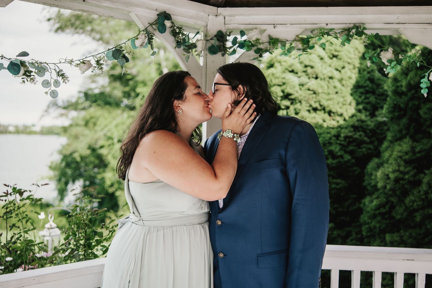 Two brides share a first kiss under a floral arch at an outdoor wedding ceremony by the water.