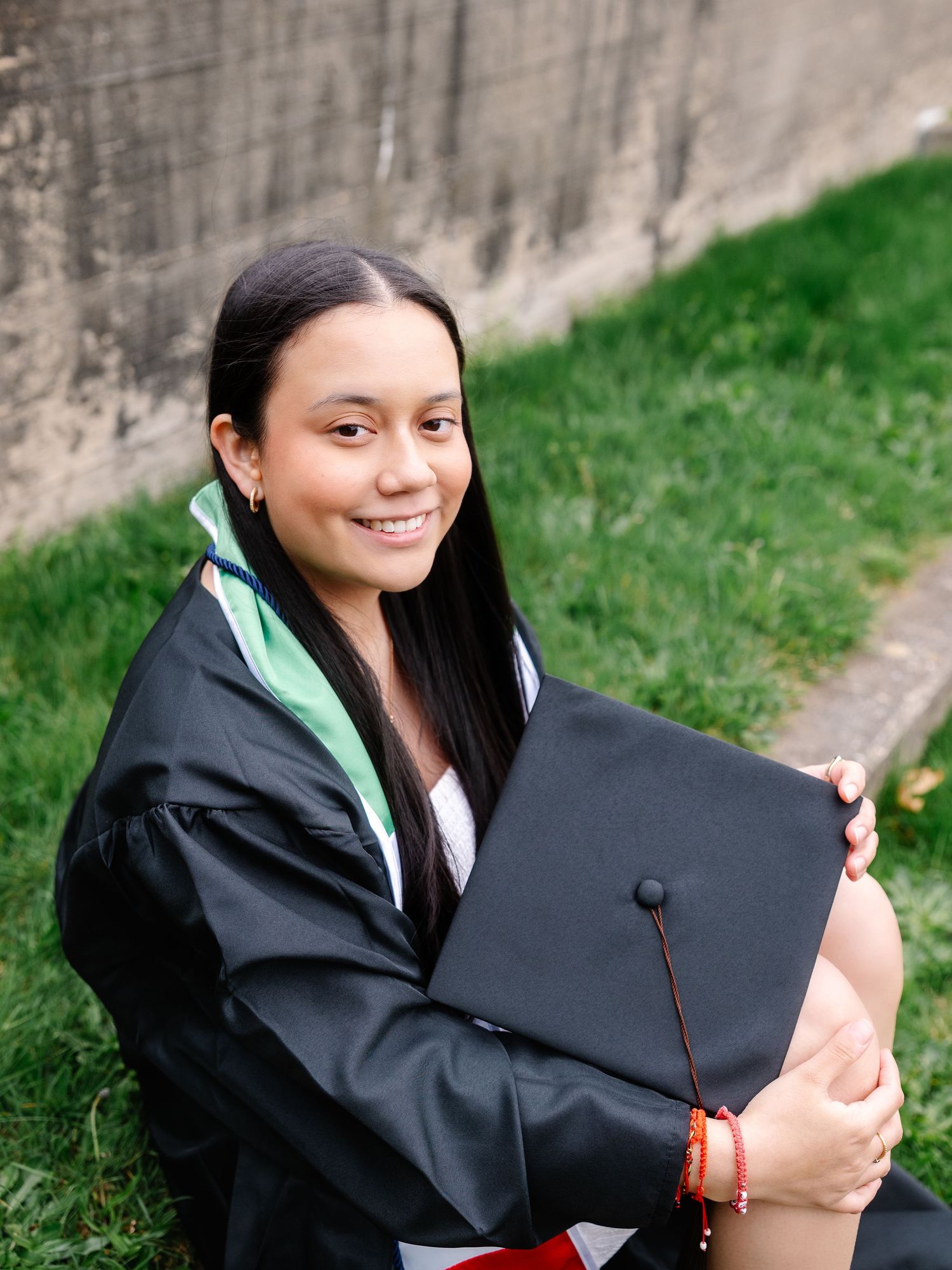 Smiling graduate hugs black mortarboard while sitting outdoors in cap and gown.