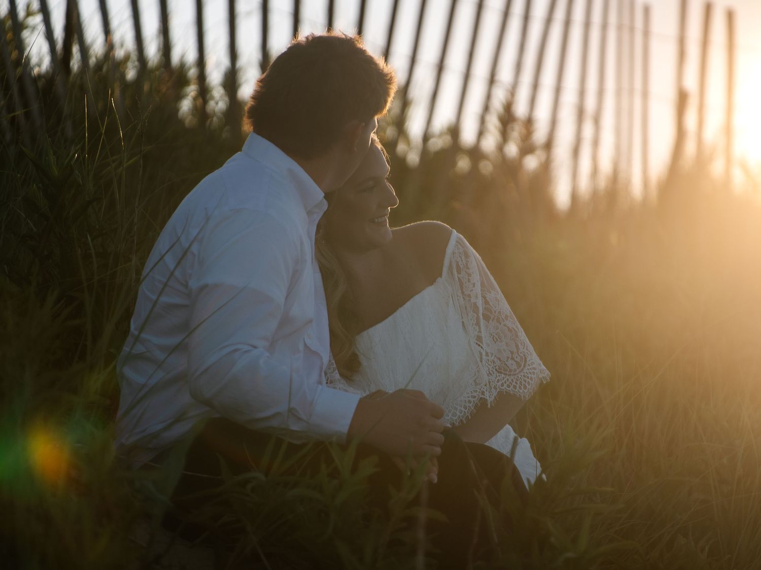 Romantic couple embracing in tall grass at golden sunset, man in white shirt kissing woman in white lace dress.