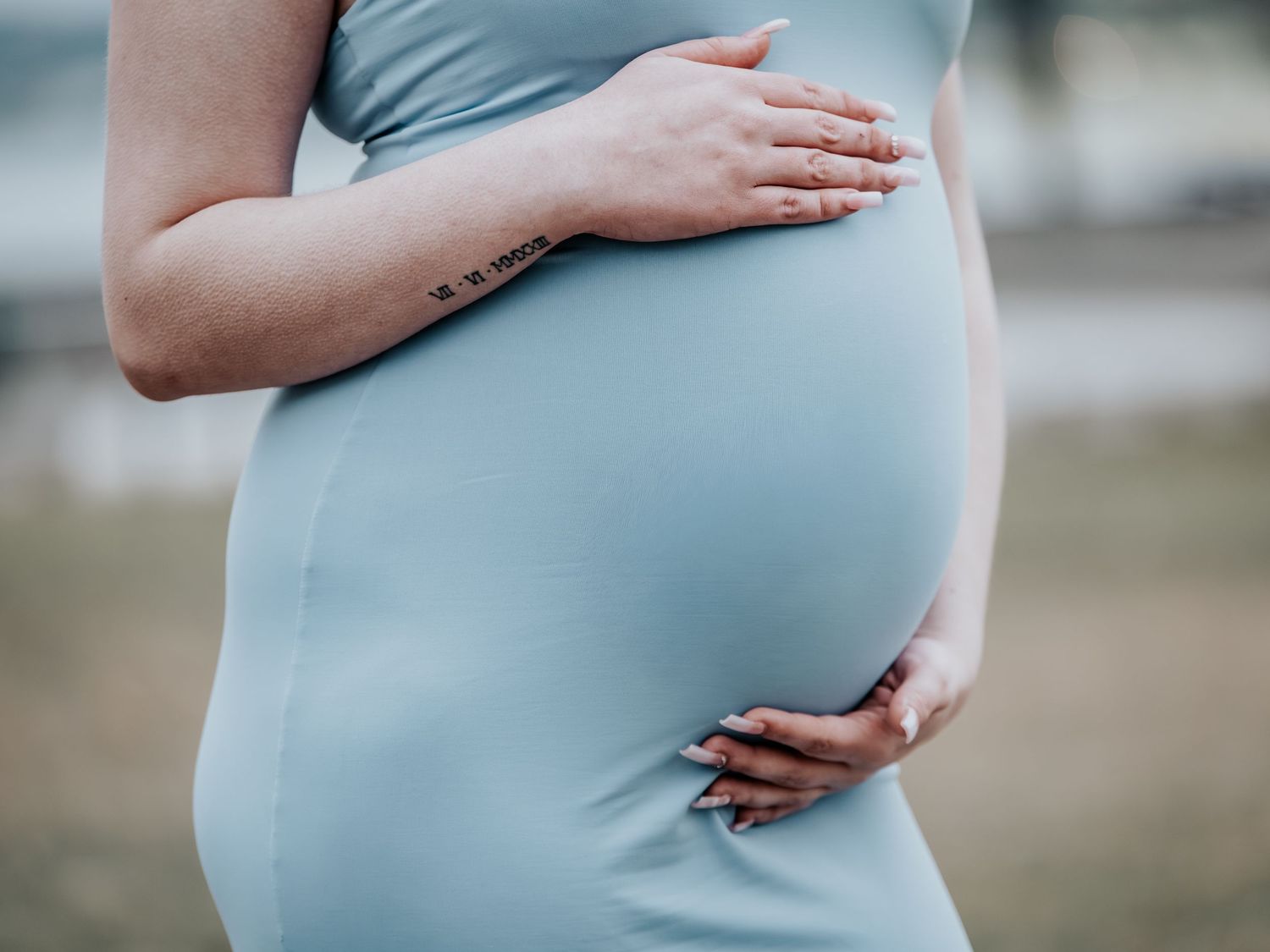 Pregnant woman in light blue dress cradling her baby bump outdoors, showcasing maternity fashion.