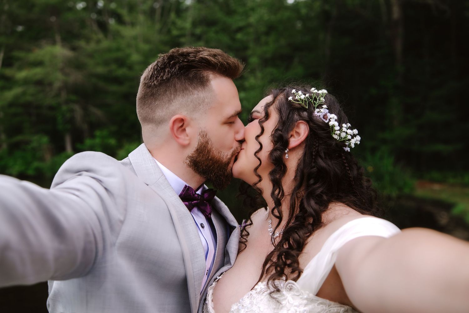 Bride and groom sharing a romantic kiss while taking a wedding selfie outdoors in a lush green forest setting.