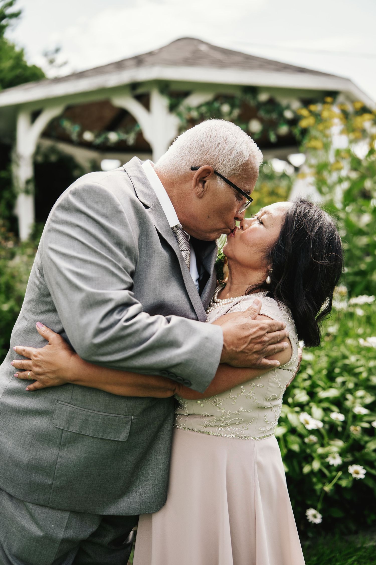 Older couple sharing a romantic kiss in a garden, man in gray suit embracing woman in blush dress near a white gazebo.