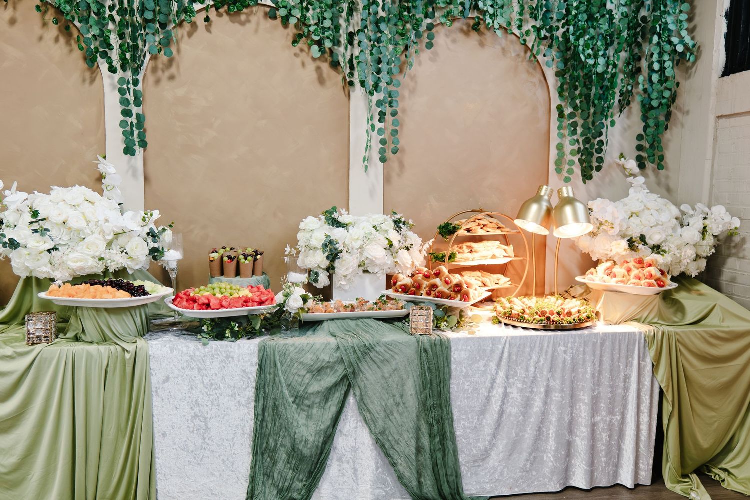 Luxurious wedding reception buffet table with green sage runner, white orchids, hanging eucalyptus backdrop, and assorted food displays.