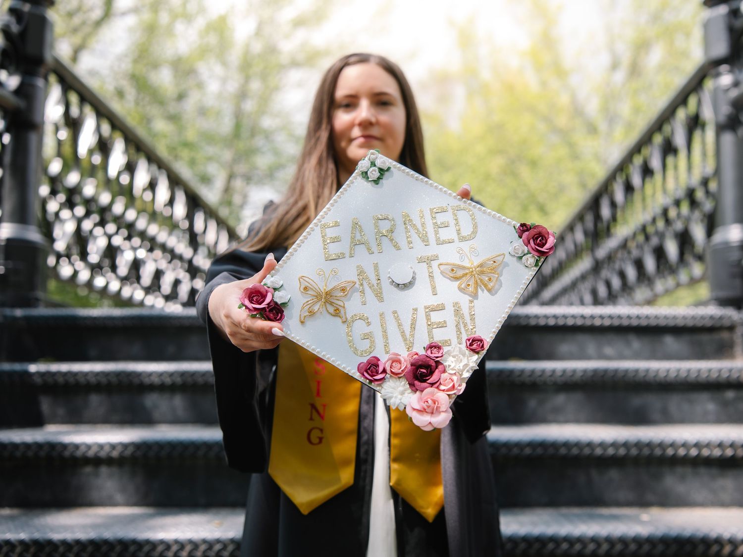 Graduate in cap and gown holding decorated mortarboard reading Earned Not Given on ornate staircase.