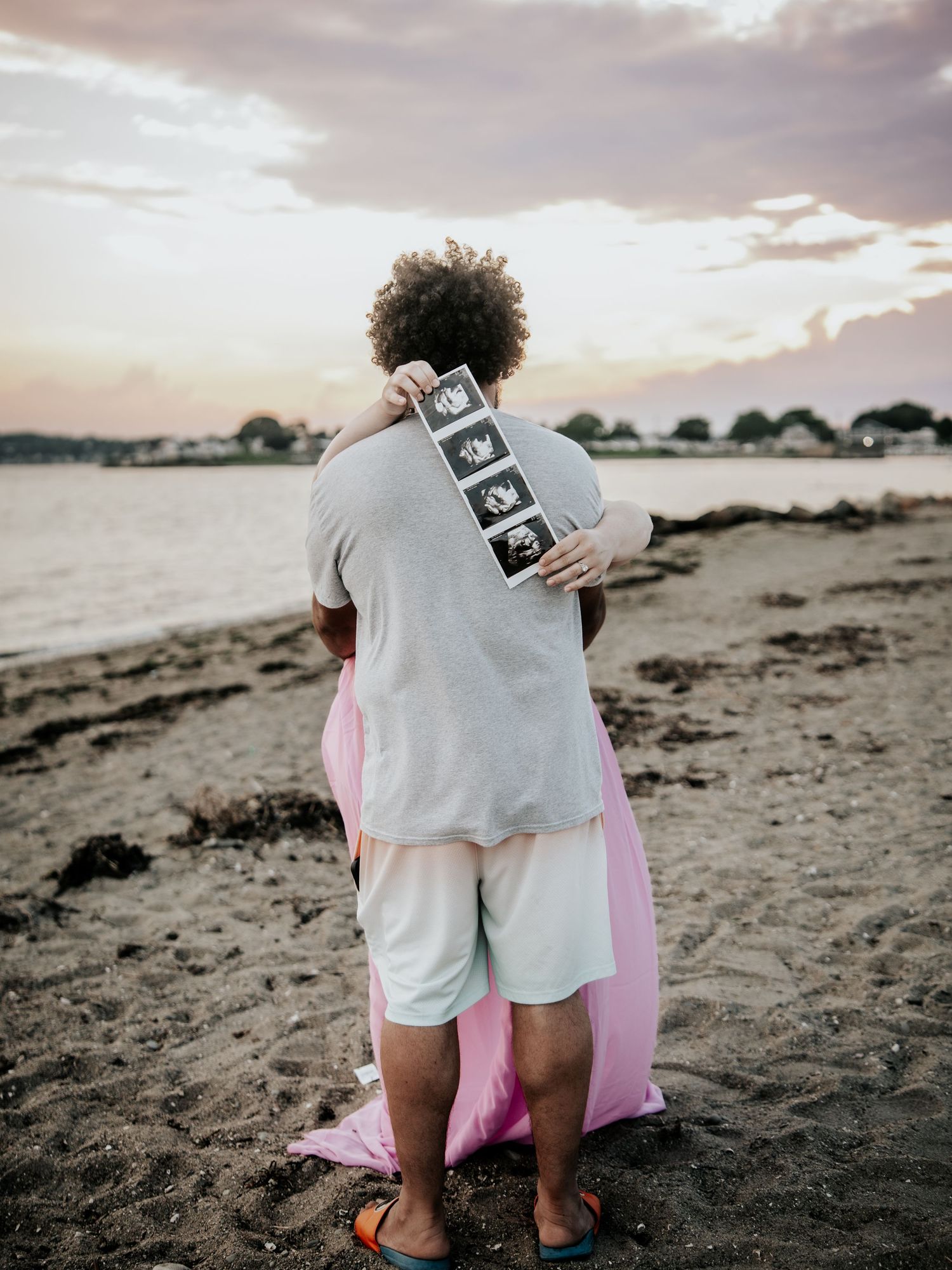 Couple embracing on a sandy beach at sunset while holding ultrasound photos.