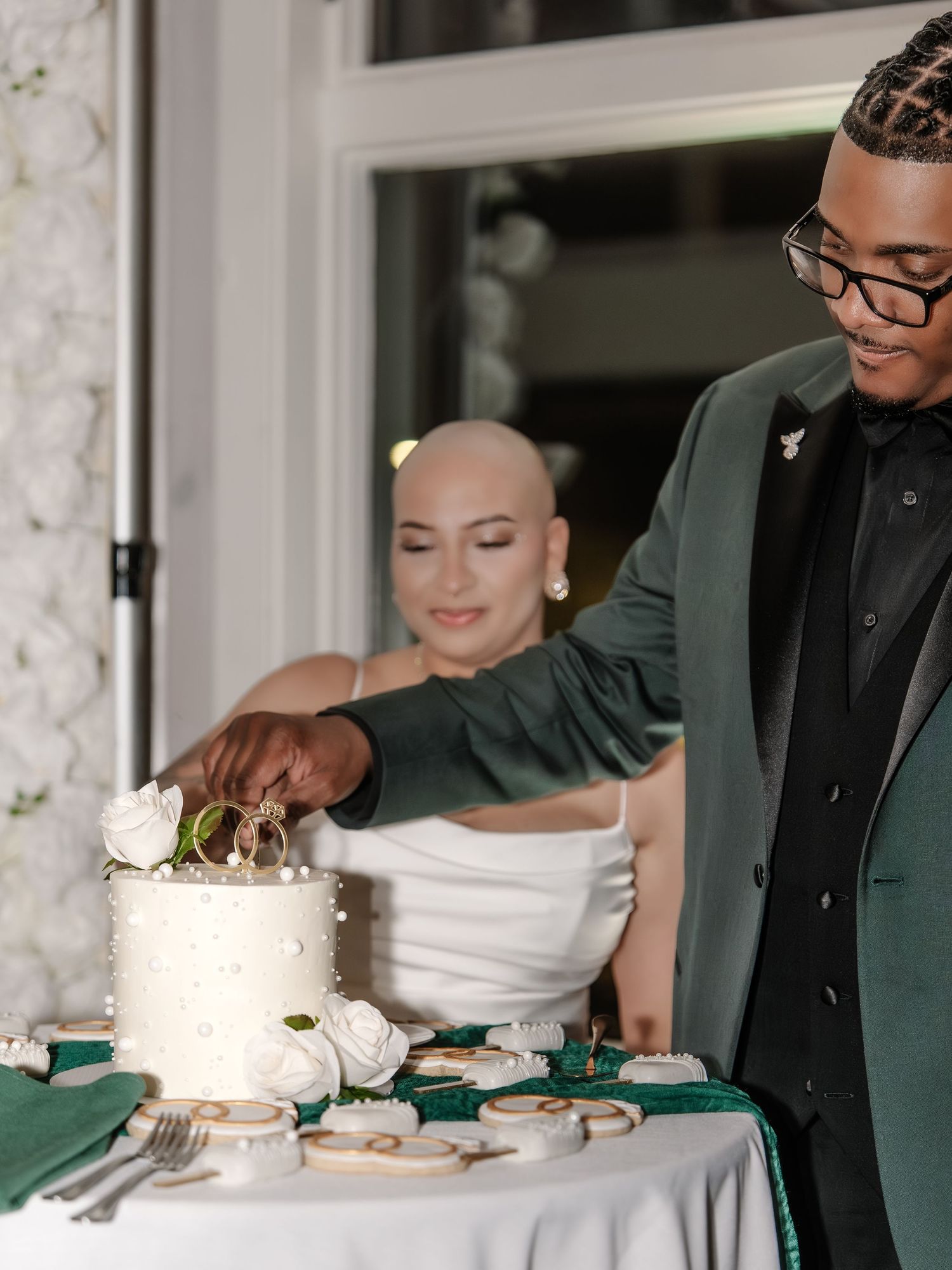 Couple cutting white pearl wedding cake, groom in green velvet suit, bride in white dress at elegant reception.