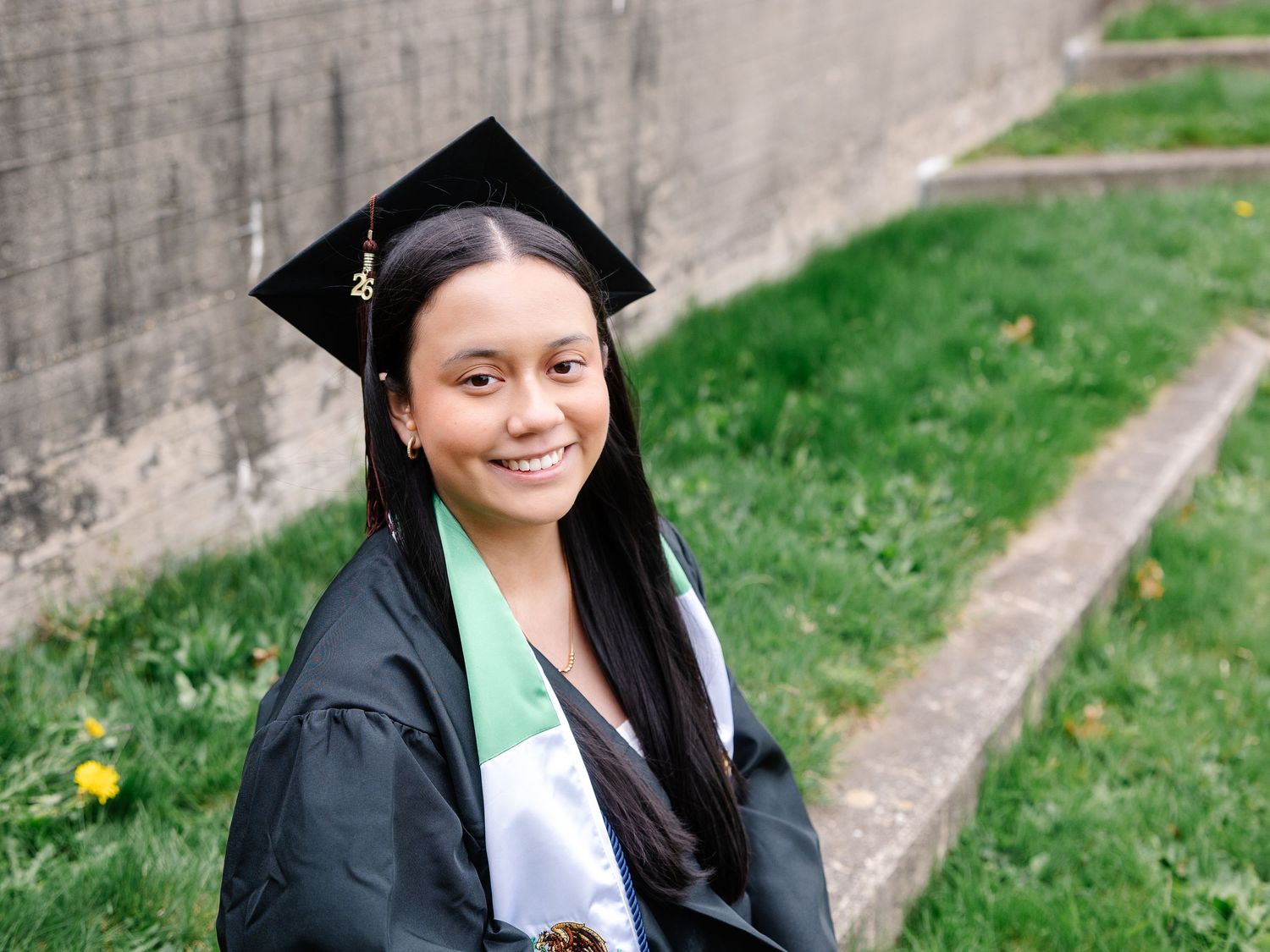 Smiling graduate in black cap and gown sits on steps with green grass background.