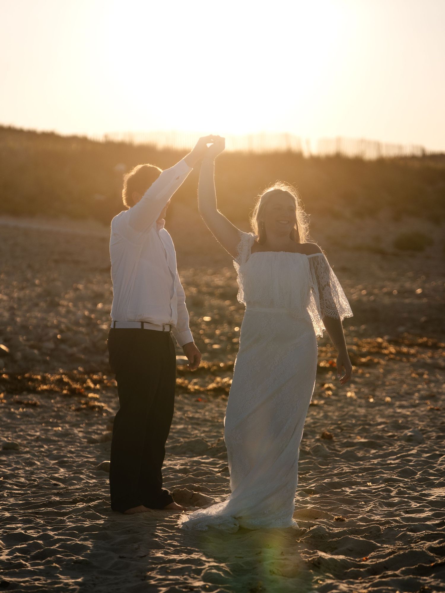 Couple holding hands raised toward golden sunset sky on sandy beach, woman in flowing white wedding dress.
