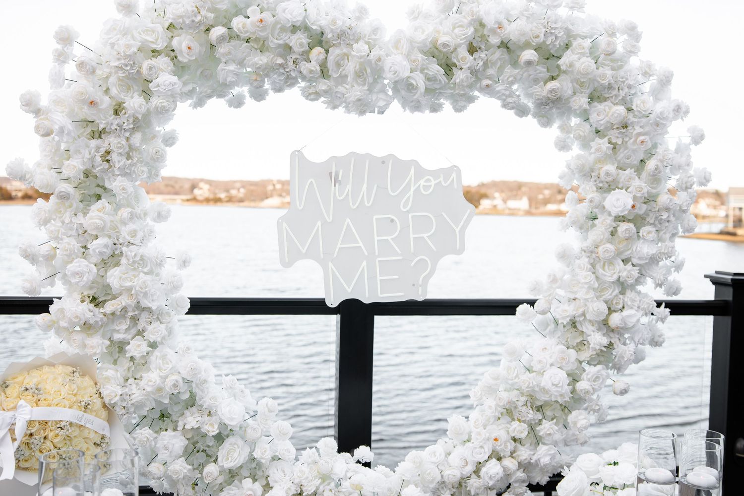 White floral heart-shaped arch with 'Will You Marry Me' sign overlooking a waterfront proposal setup.