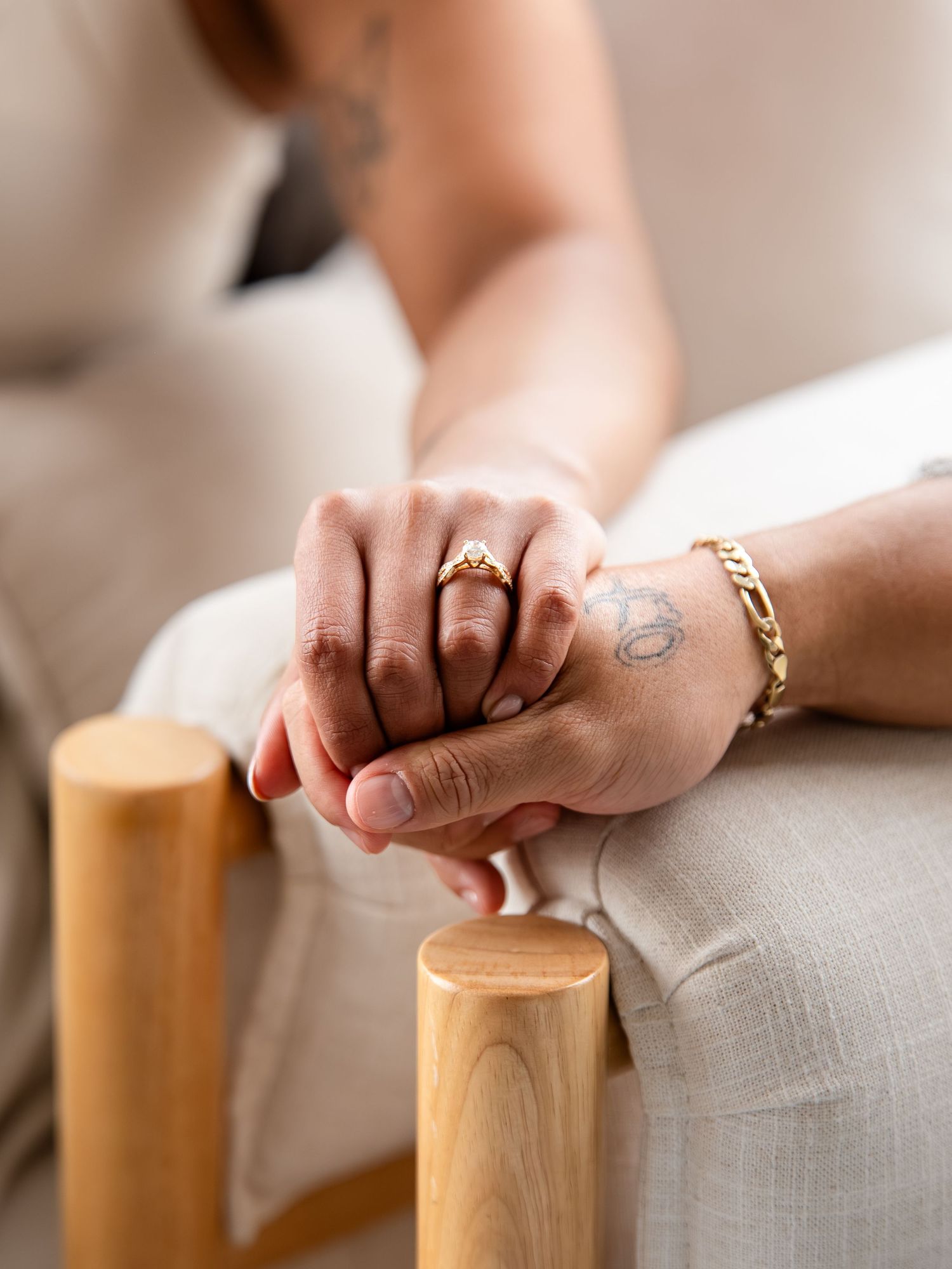 Two hands intertwined, one wearing a gold ring and the other a gold bracelet with a tattoo, resting on a chair.