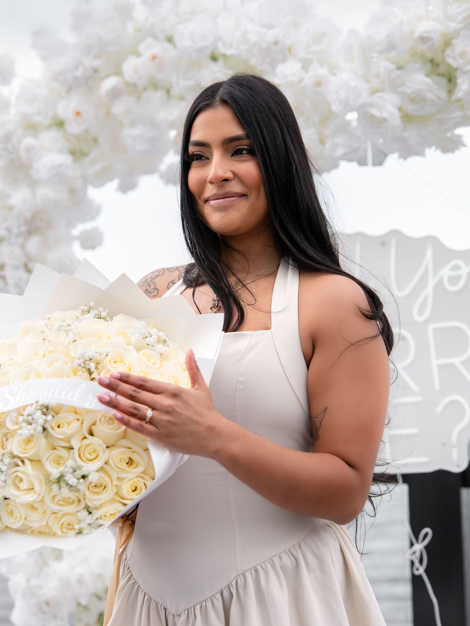 Smiling woman in white dress holding a bouquet of white roses at an outdoor proposal setup.