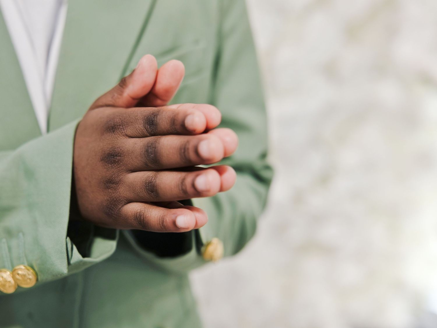 Close-up detail of clasped hands wearing a mint green suit jacket with decorative gold buttons at a formal event.