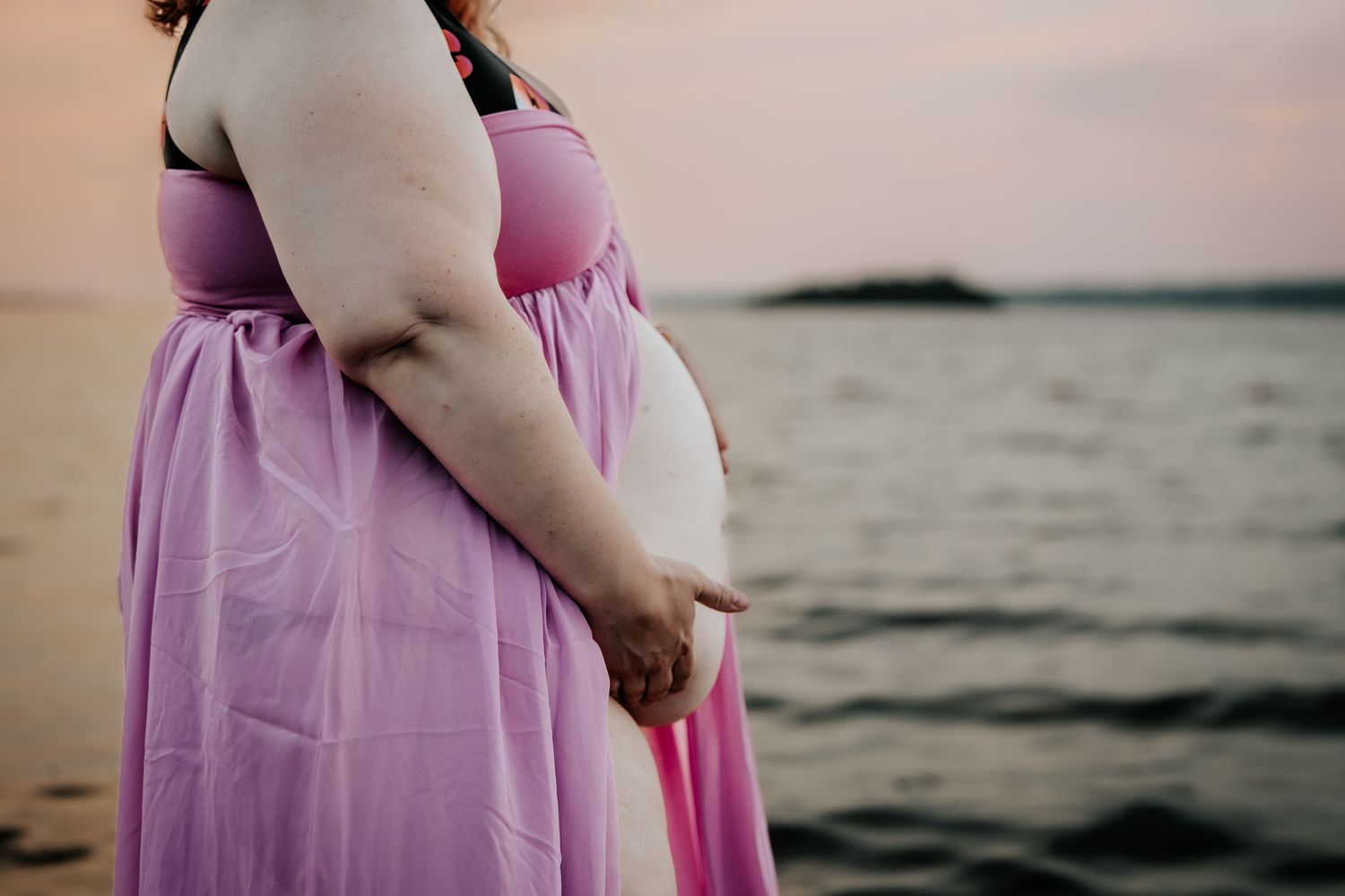Pregnant woman in pink dress standing by the ocean at sunset, showing baby bump.