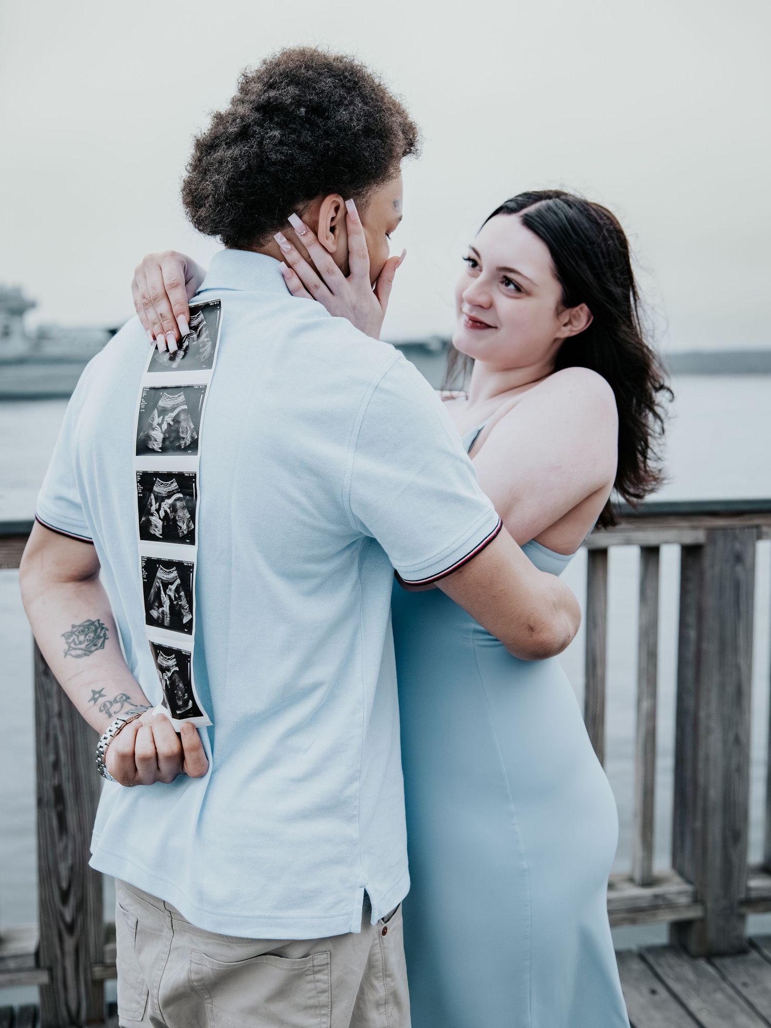 Couple sharing a tender moment on a wooden dock, man holding sonogram photos on his back, pregnancy announcement.