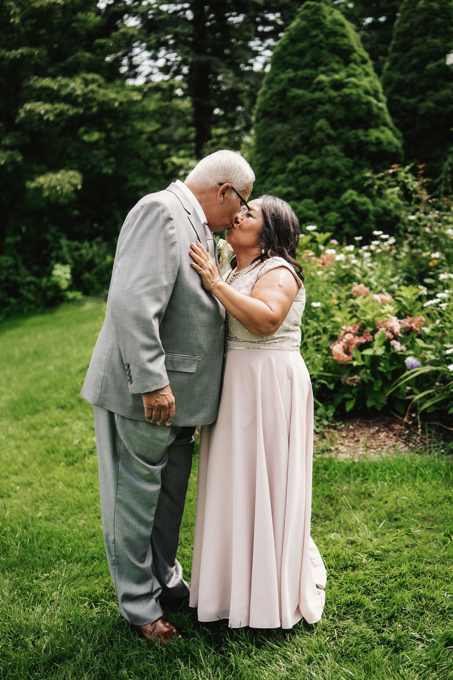 Elderly couple sharing a romantic kiss in a lush garden, dressed in formal wedding attire, gray suit and blush gown.
