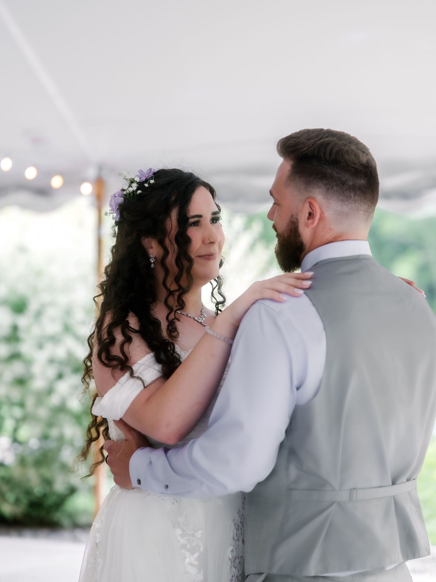 Bride and groom share first dance under white tent, bride wearing floral crown and lace gown.