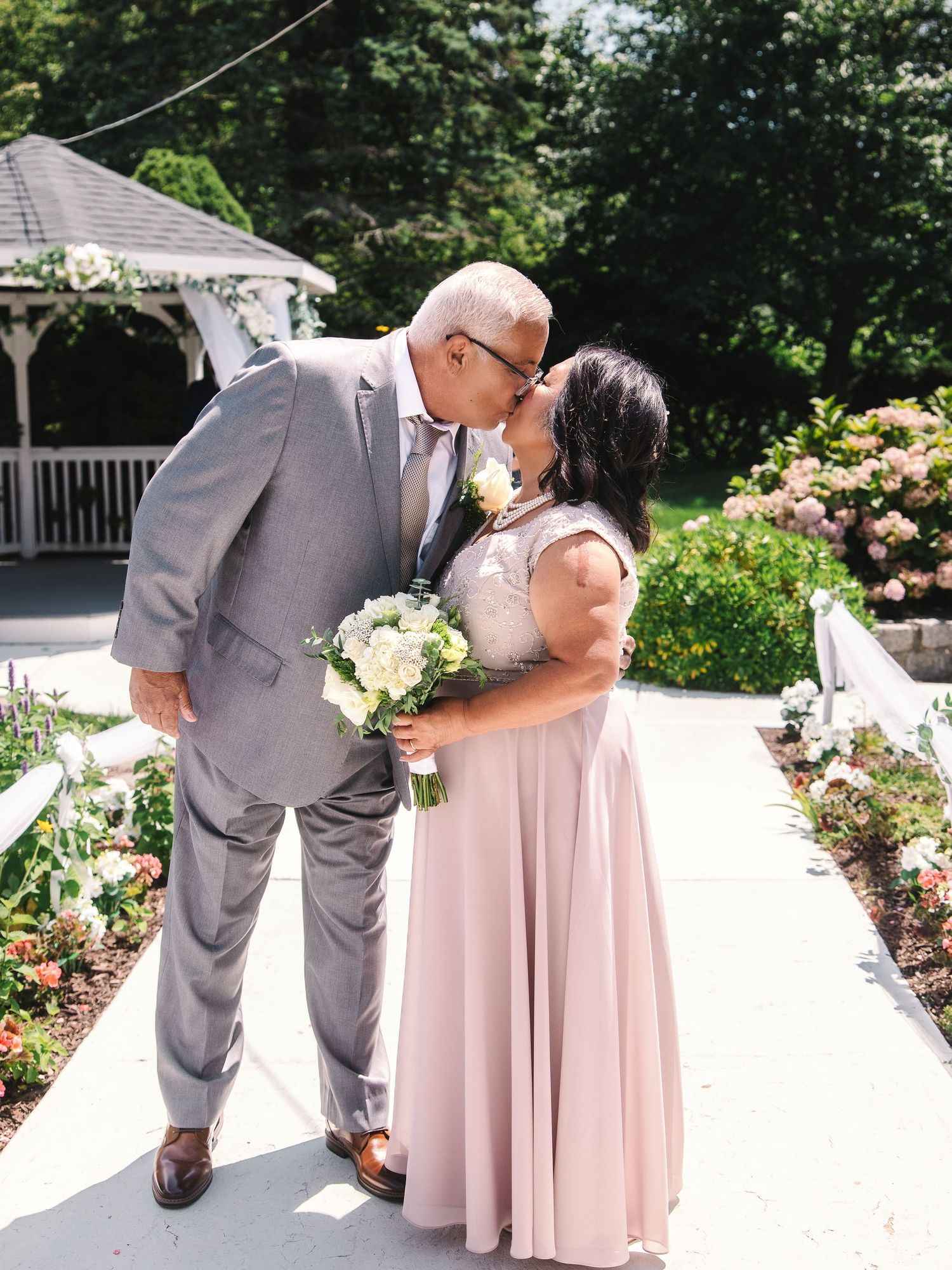 Couple sharing a kiss in wedding attire near a gazebo, surrounded by blooming flowers on a sunny day.