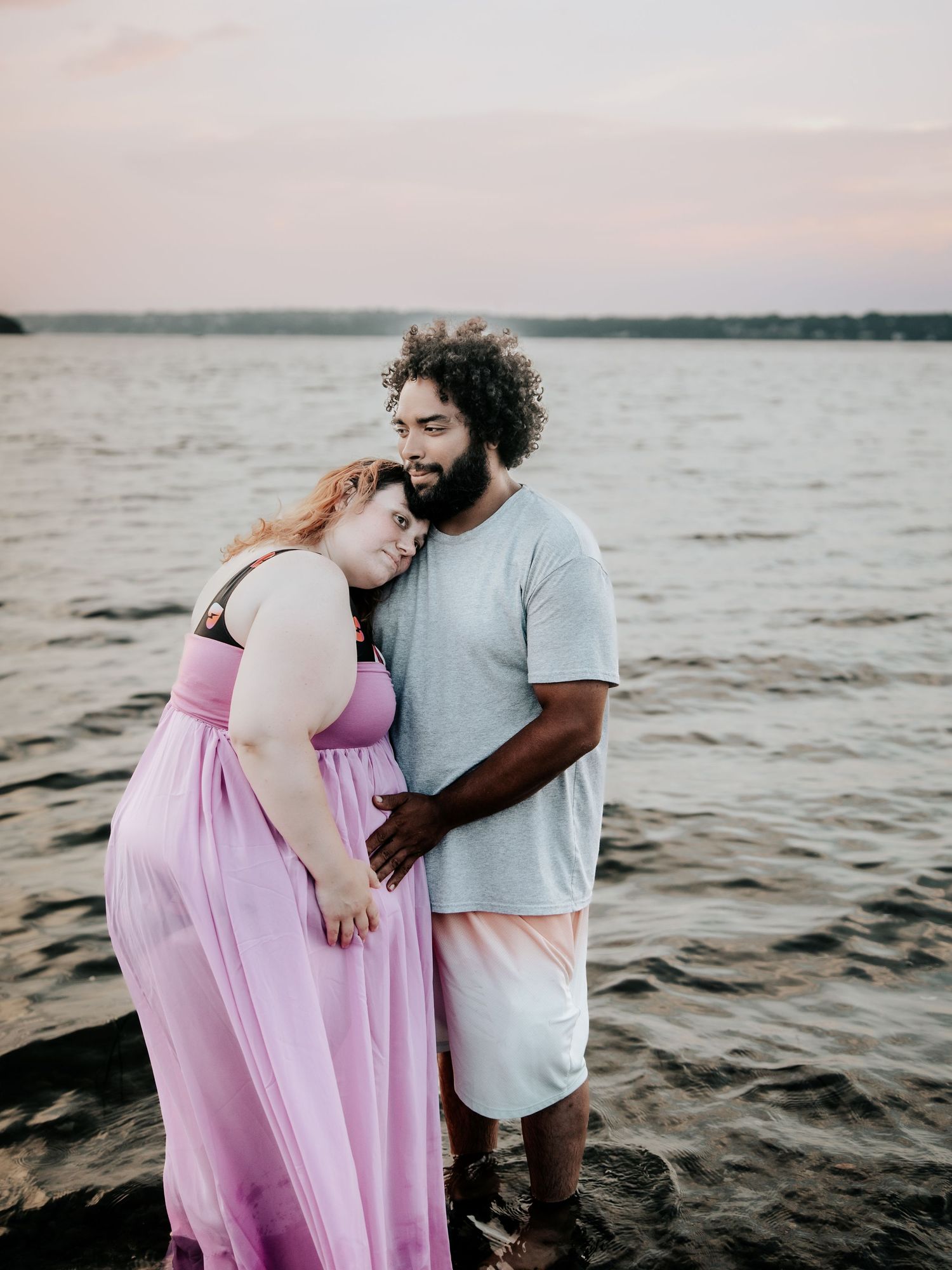 A couple shares a tender moment at sunset by the water, woman in pink gown leaning on mans shoulder.