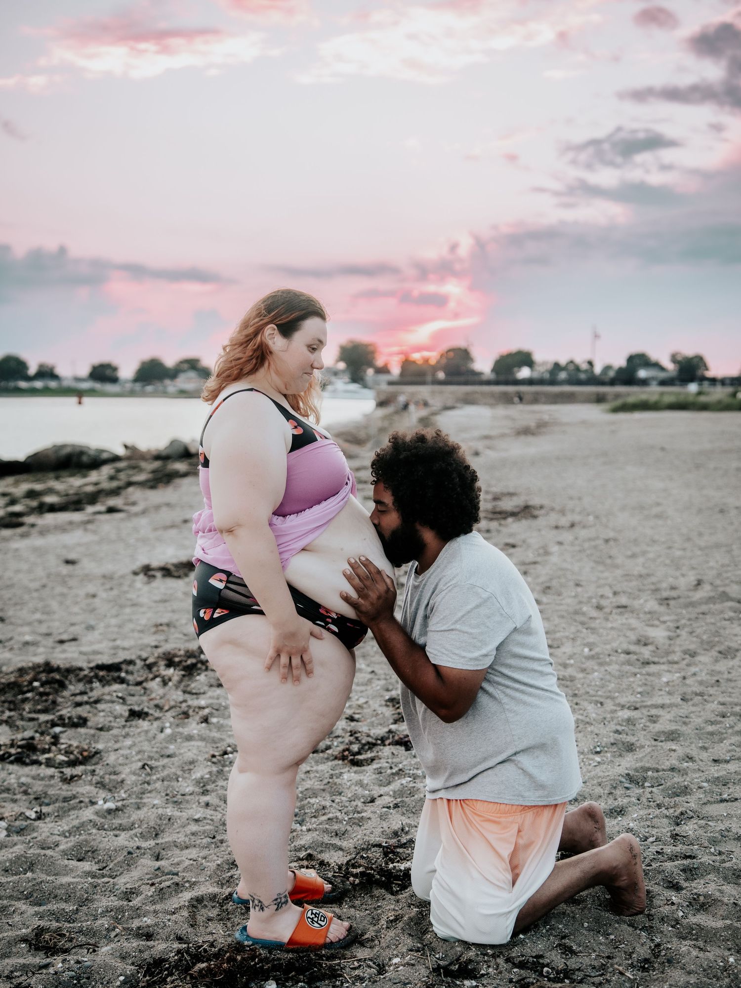 Pregnant woman in pink bikini top on beach at sunset with partner kneeling and touching her belly.
