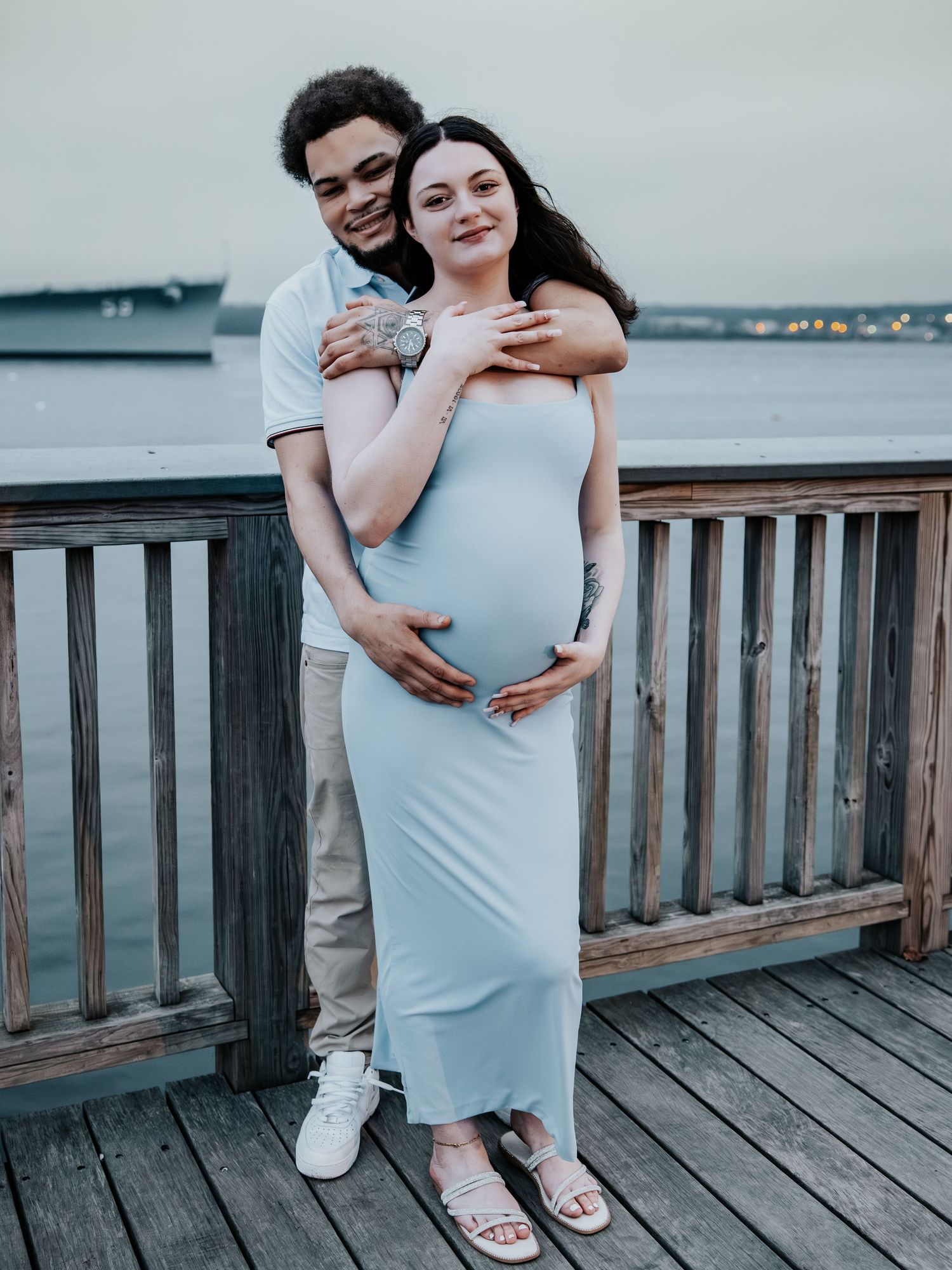 Expecting couple embracing on waterfront pier, woman in light blue maternity dress showing baby bump.