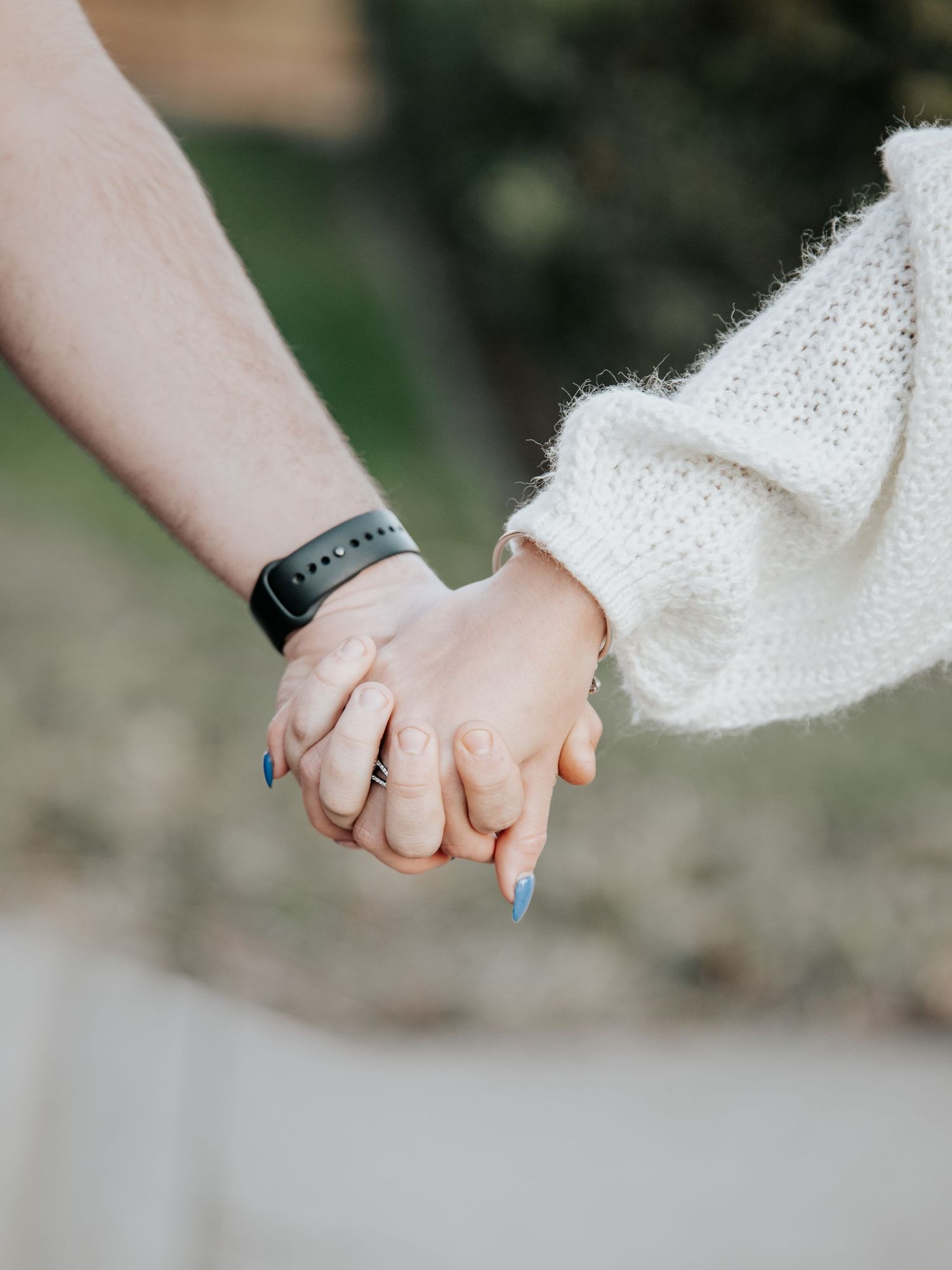 Two people holding hands outdoors, one wearing a black fitness tracker, the other in a white knit sweater with blue nails.