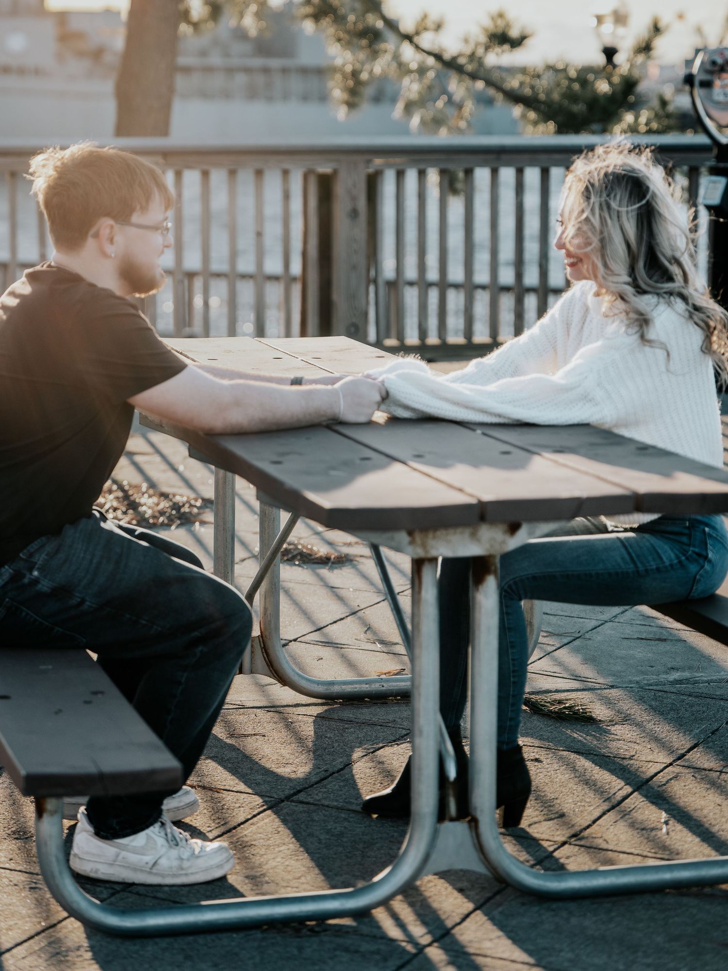Couple holding hands across a picnic table outdoors at sunset during engagement photo session.
