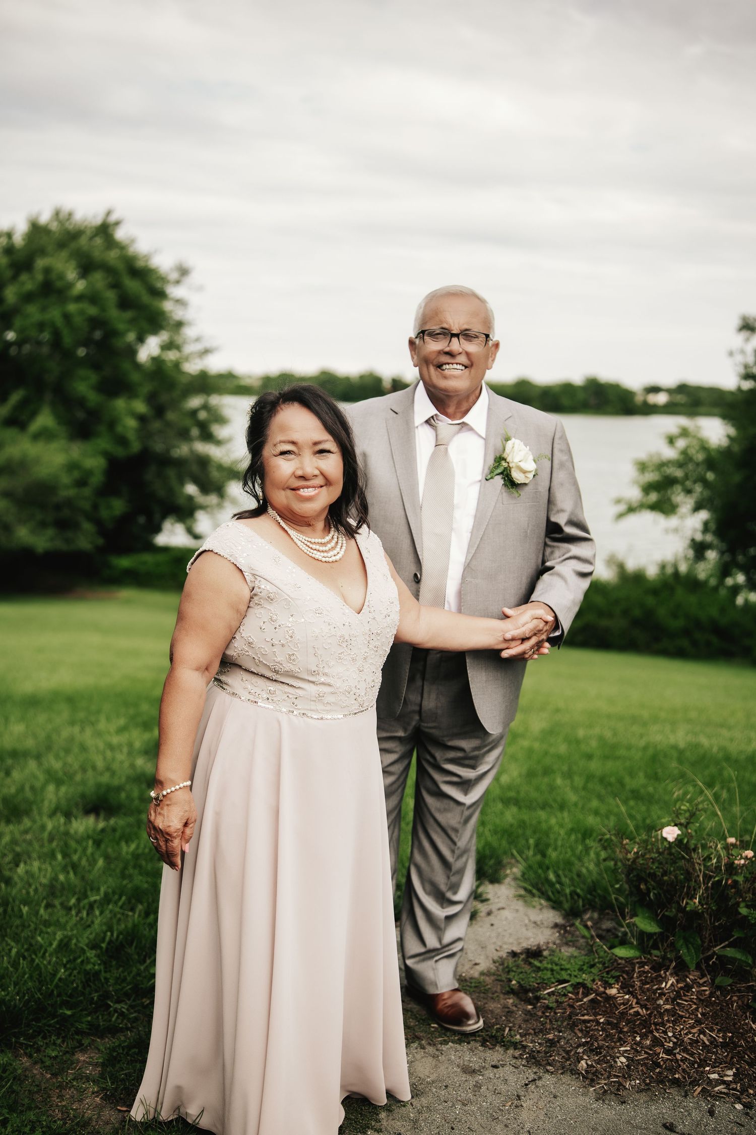 Elegant couple posing outdoors near a lake, wearing formal wedding attire including a gray suit and blush gown.