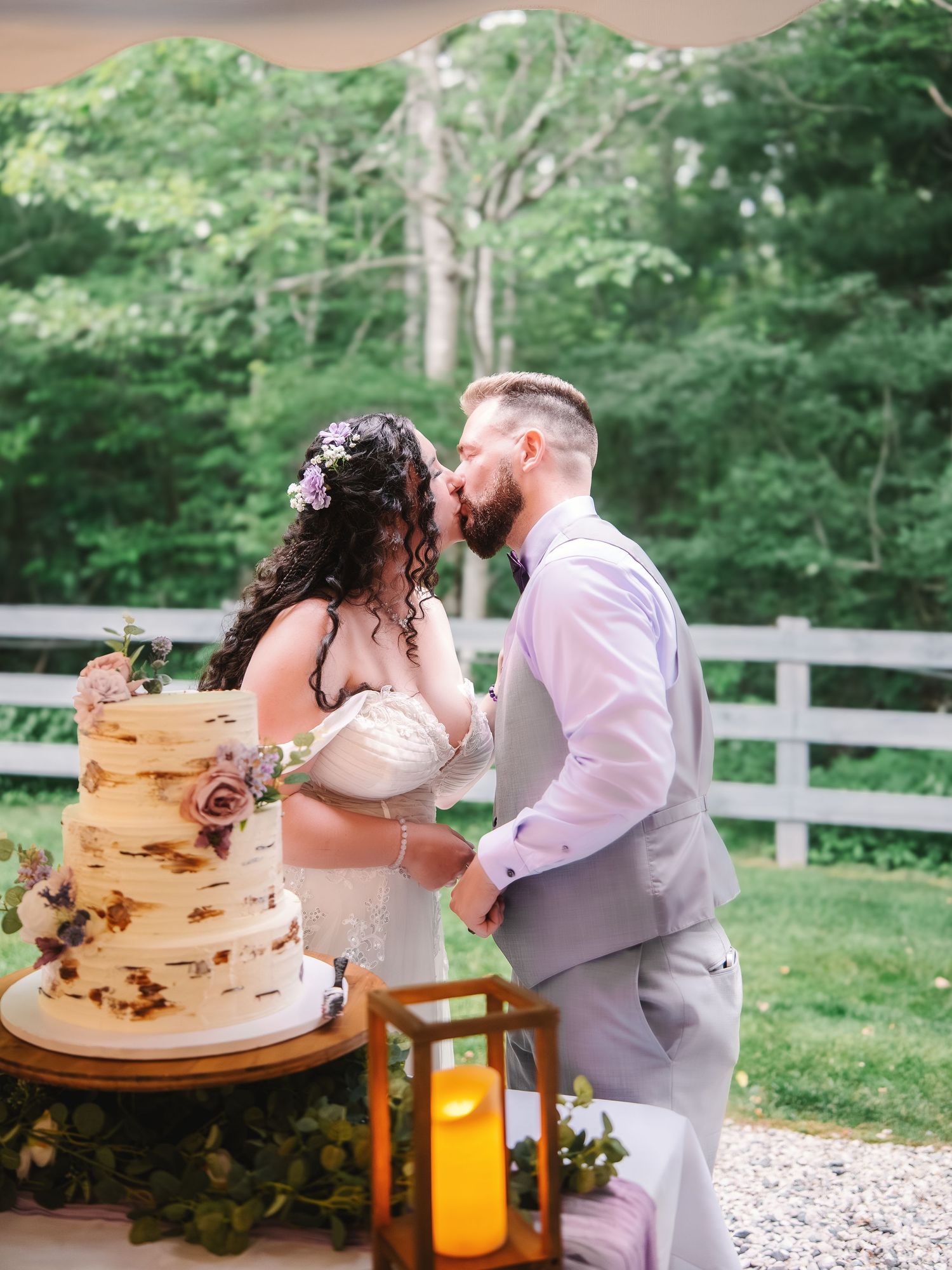 Bride and groom sharing a kiss beside a rustic three-tier wedding cake outdoors with a white fence backdrop.