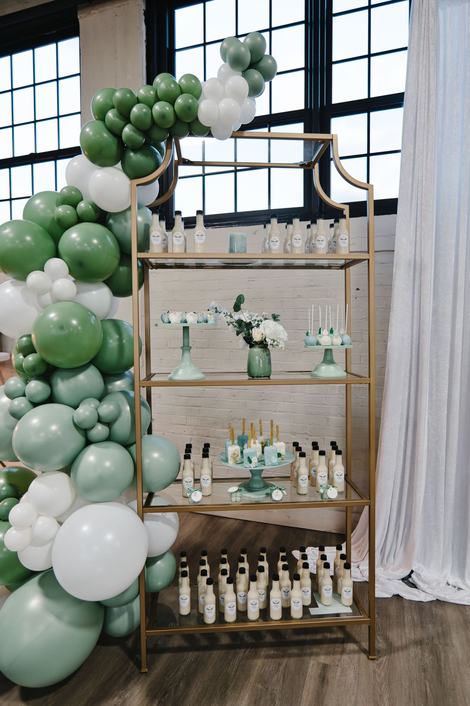 Gold display shelf with baptism favors and white flowers surrounded by sage green balloon garland.
