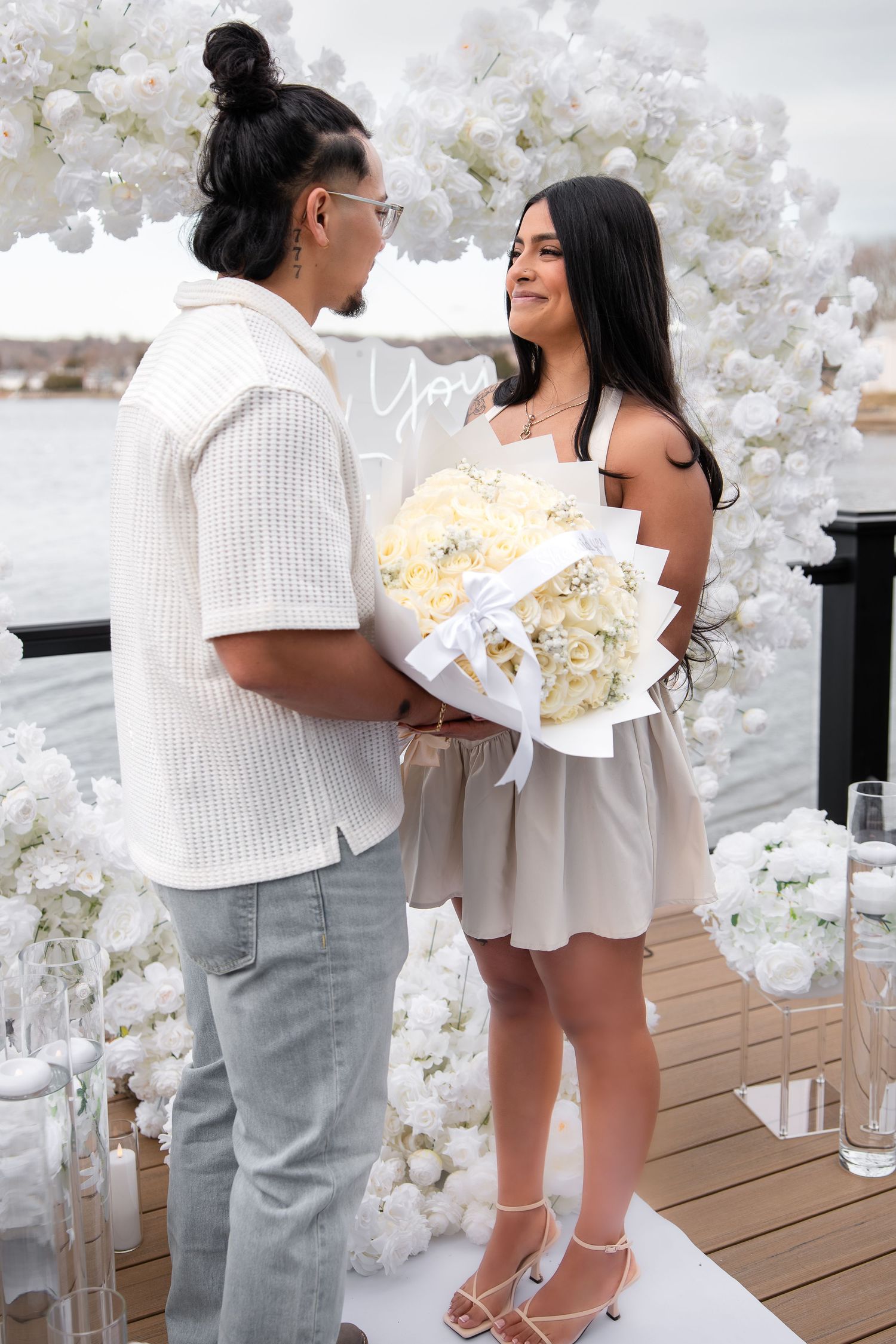 Couple stands together at waterfront proposal setup with white flowers and bouquet.