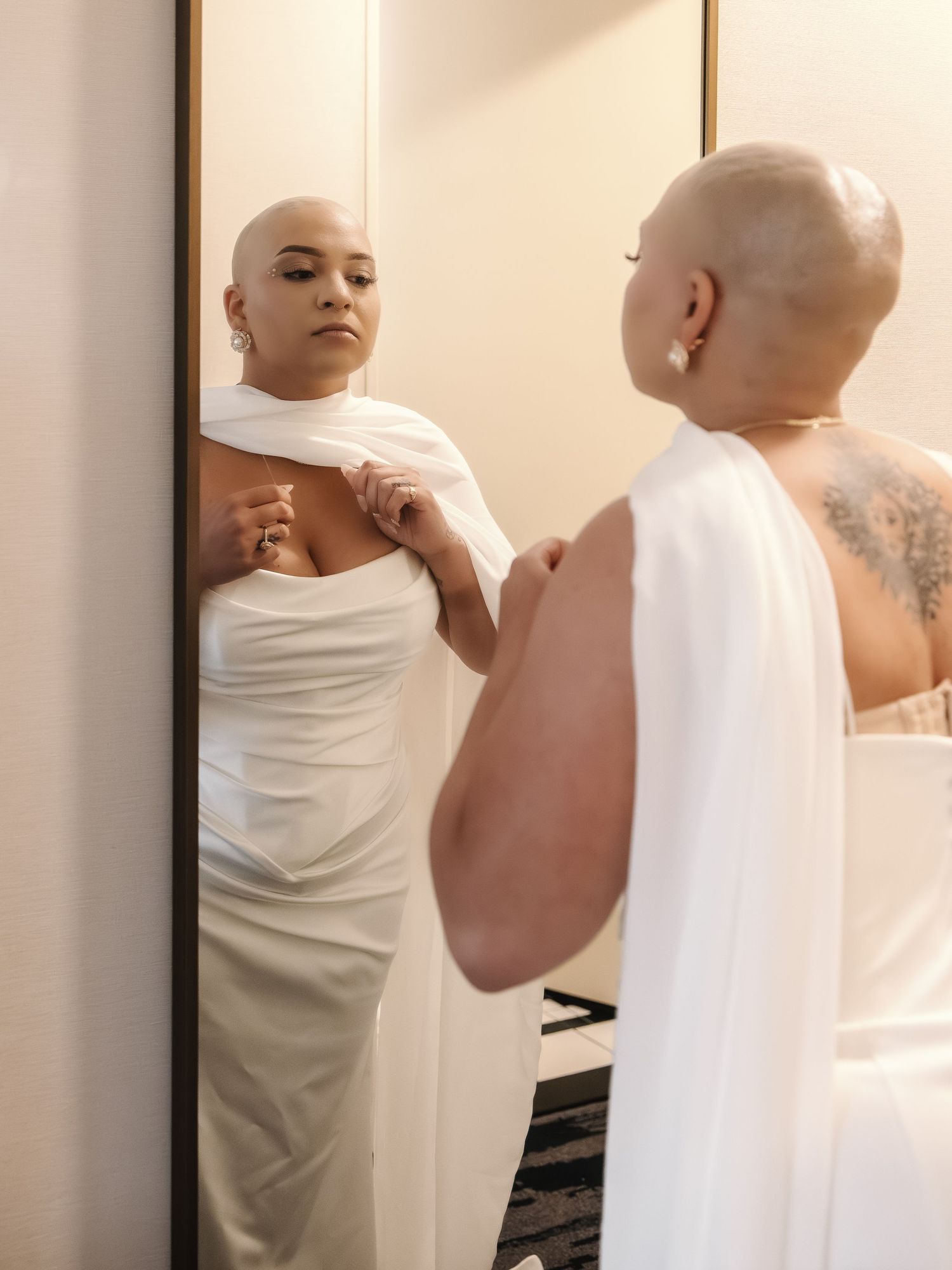 Bald woman in white strapless gown adjusting her dress while looking at her reflection in a full-length mirror.