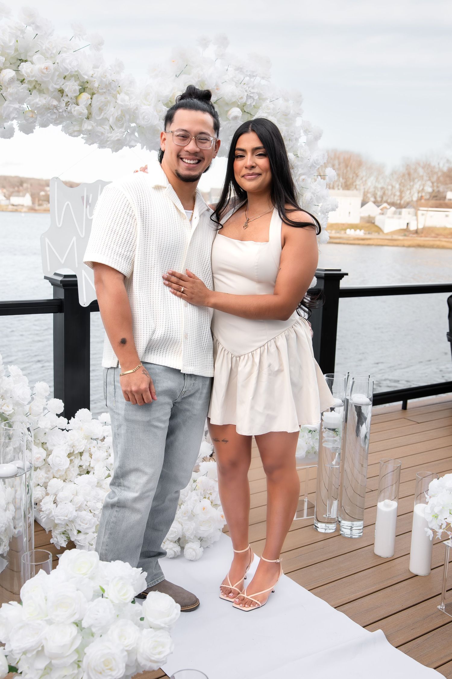 Couple in white outfits posing on a waterfront deck surrounded by white floral arrangements and candles.