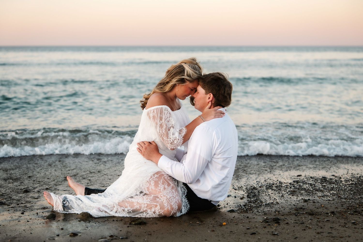 Newlyweds sitting together on wet sand sharing a kiss with gentle ocean waves behind them.