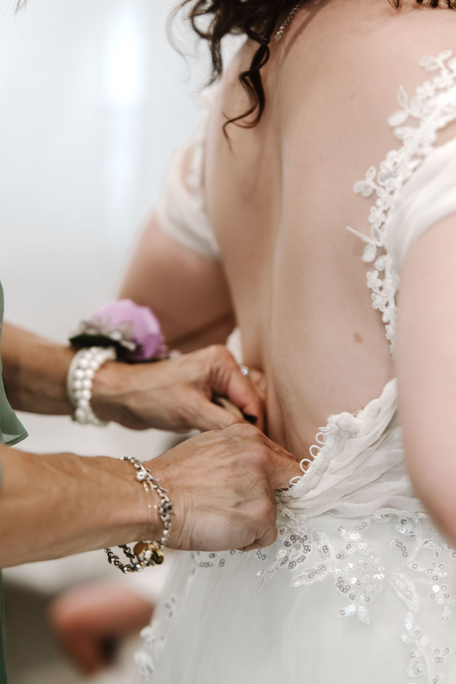 Bride getting her lace wedding dress buttoned up from behind by a helper wearing pearl bracelets.