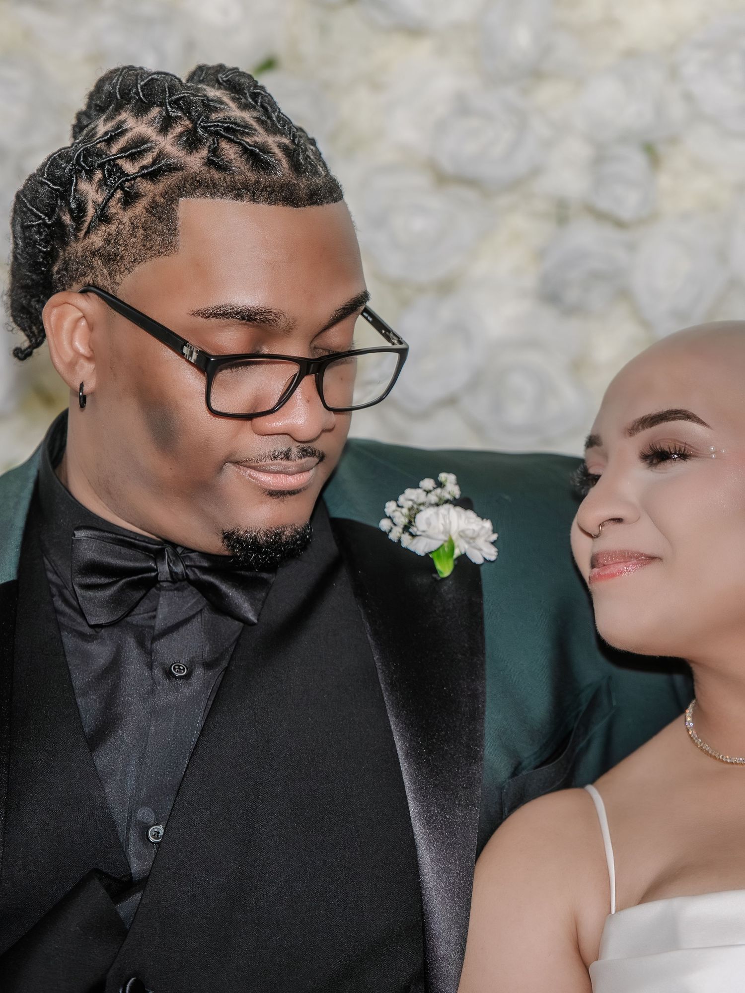 A couple in formal attire poses together, the man in a green tuxedo with braids and glasses, the woman in a white dress.