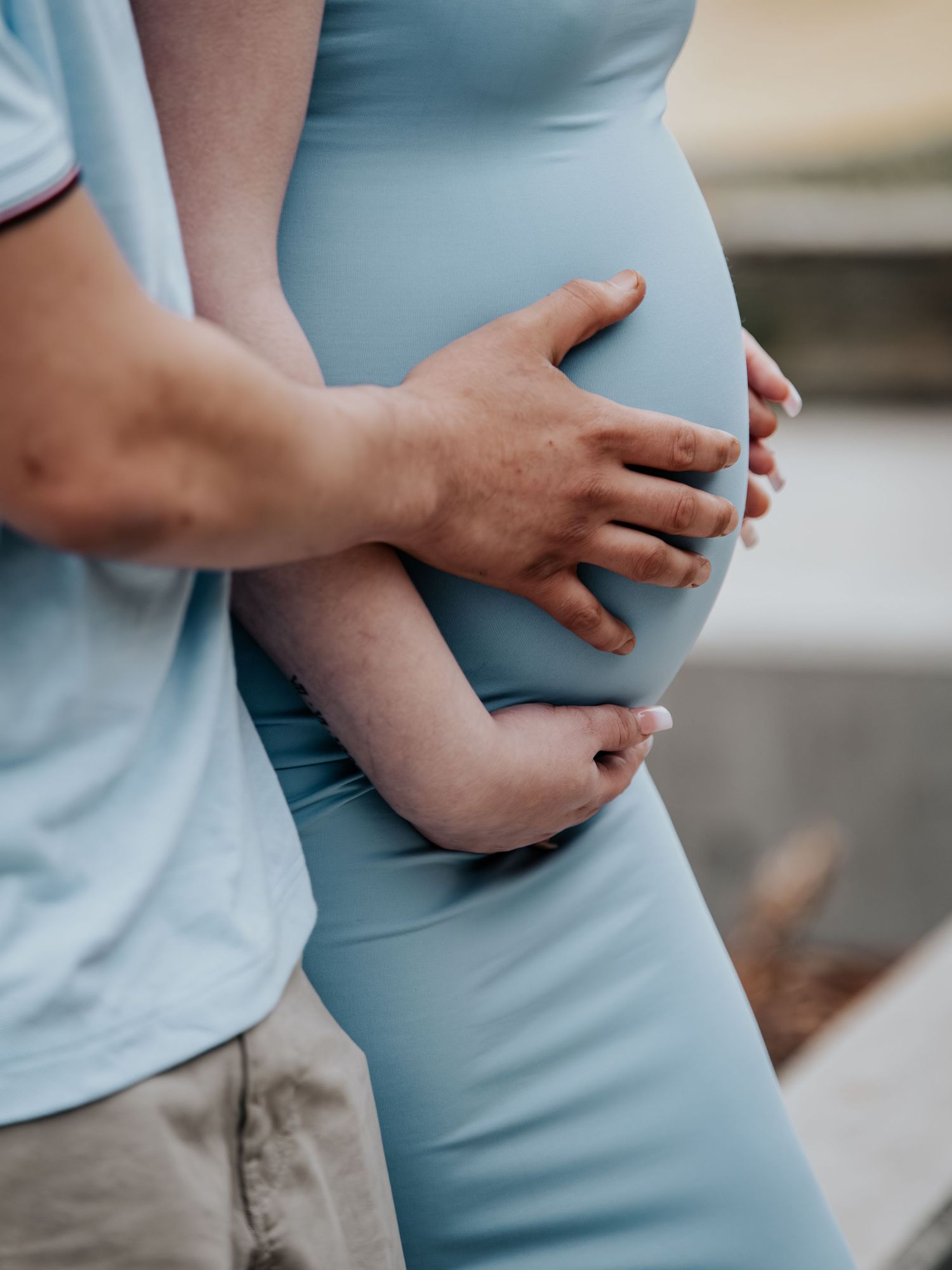 A man tenderly embraces a pregnant woman in a blue dress, cradling her baby bump with both hands.
