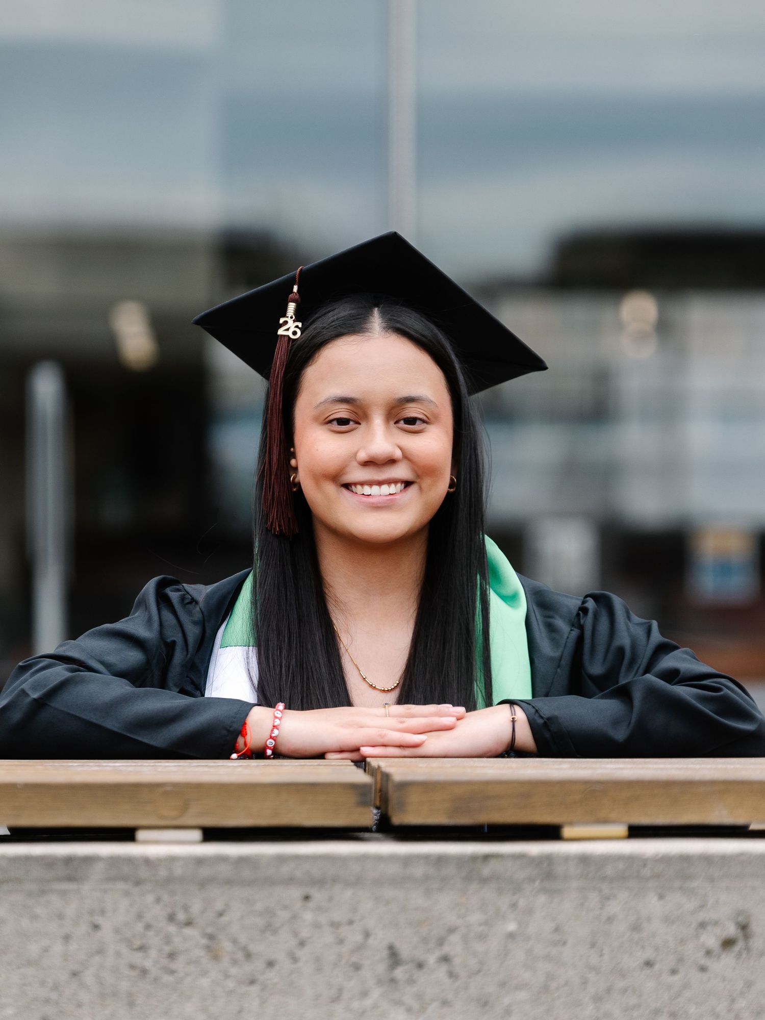 Young woman in graduation cap and gown leans on railing smiling at camera.