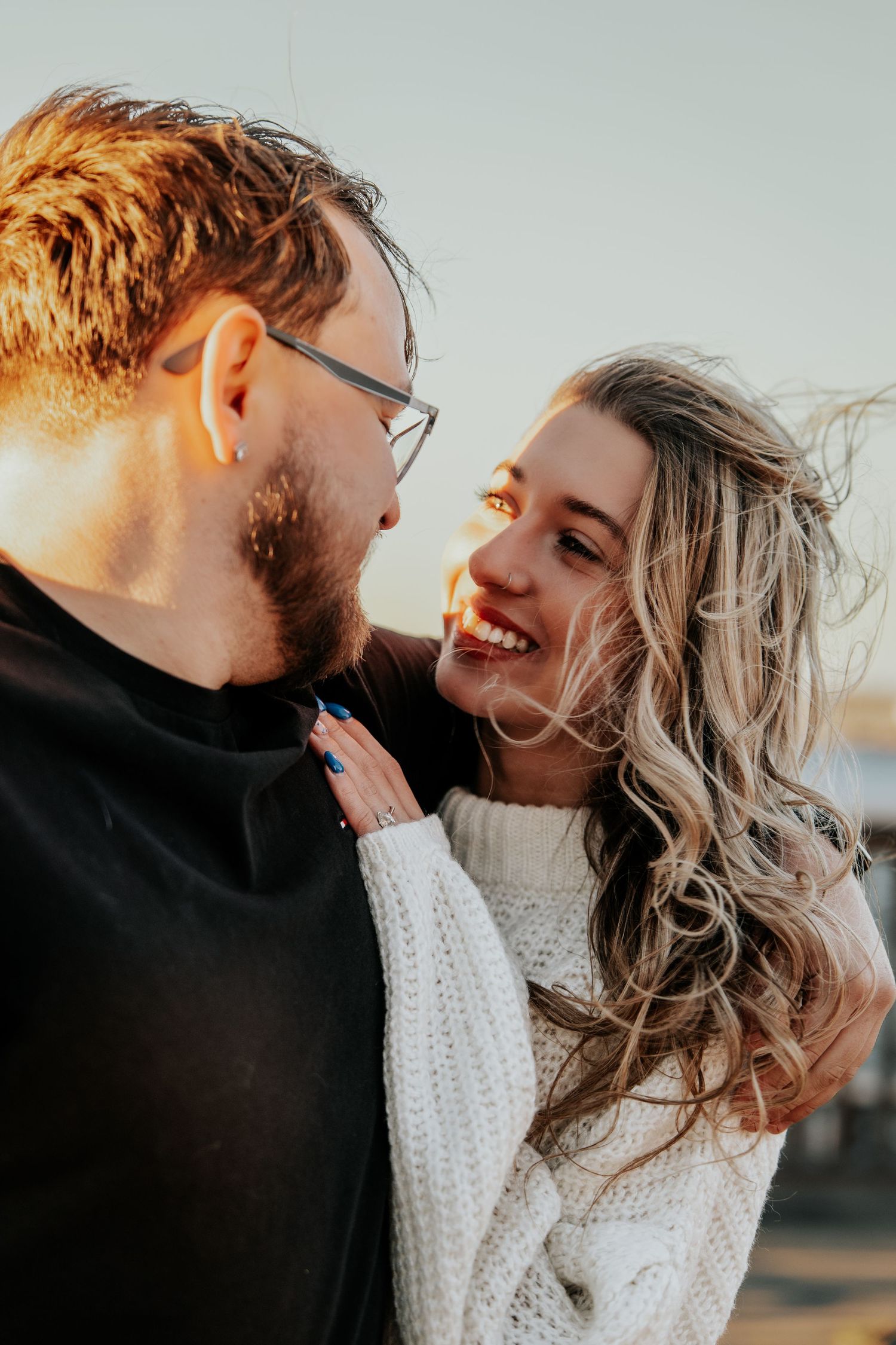 Laughing woman in white sweater embraces man in black shirt during golden hour engagement shoot.