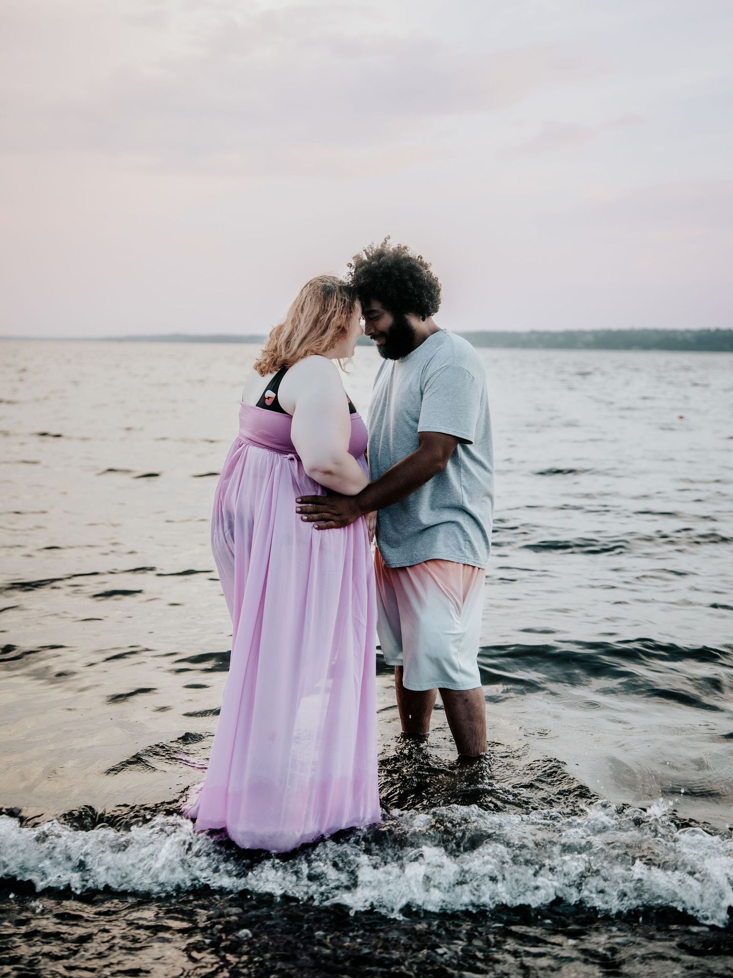 Couple shares intimate moment standing in ocean waves at sunset, woman in flowing lavender gown.