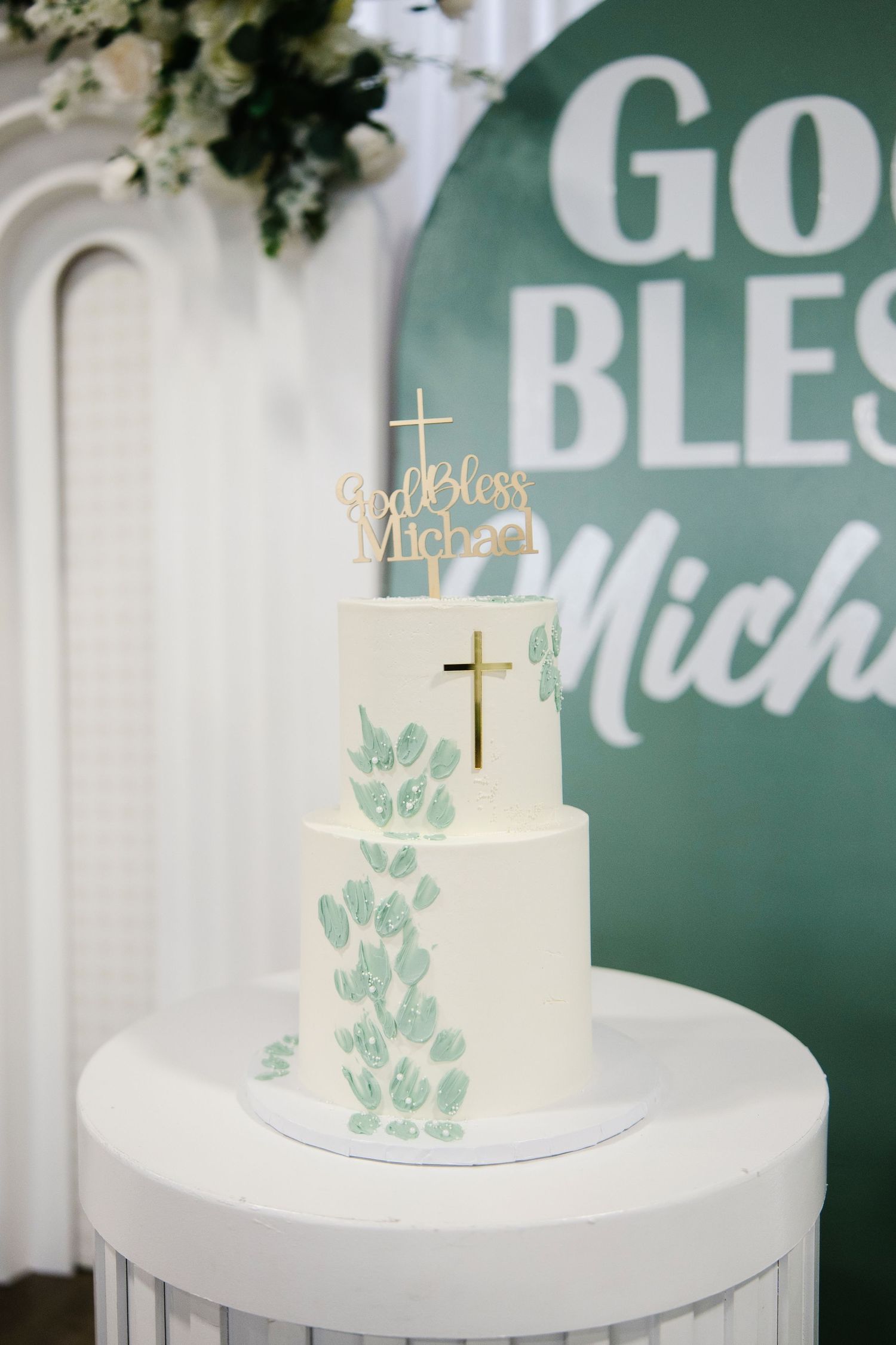 Three-tier white baptism cake with gold cross topper and green leaf design reading God Bless Michael.