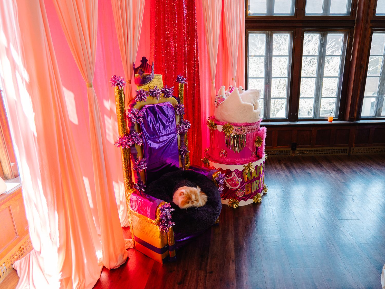 Royal purple throne chair beside a decorative birthday cake prop with pink draped curtains backdrop.