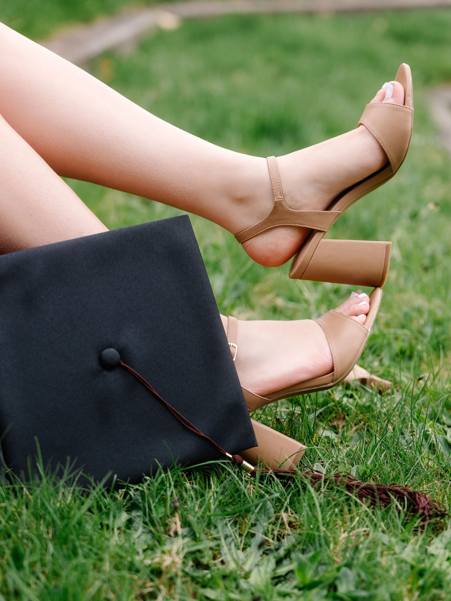Graduate sitting on grass wearing nude heeled sandals next to black graduation cap.