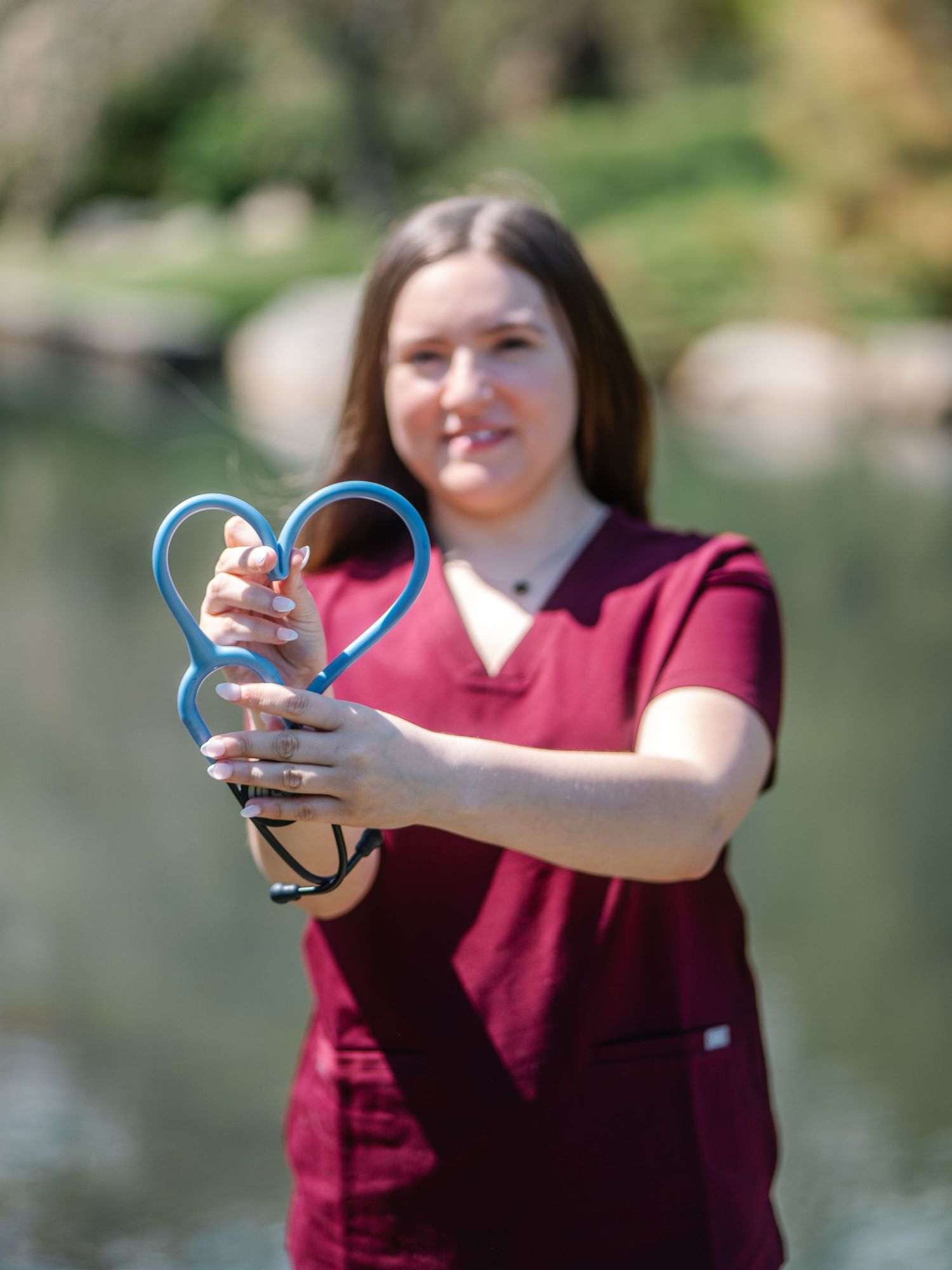 Nursing student in maroon scrubs holding blue stethoscope in heart shape near a sunlit lake.