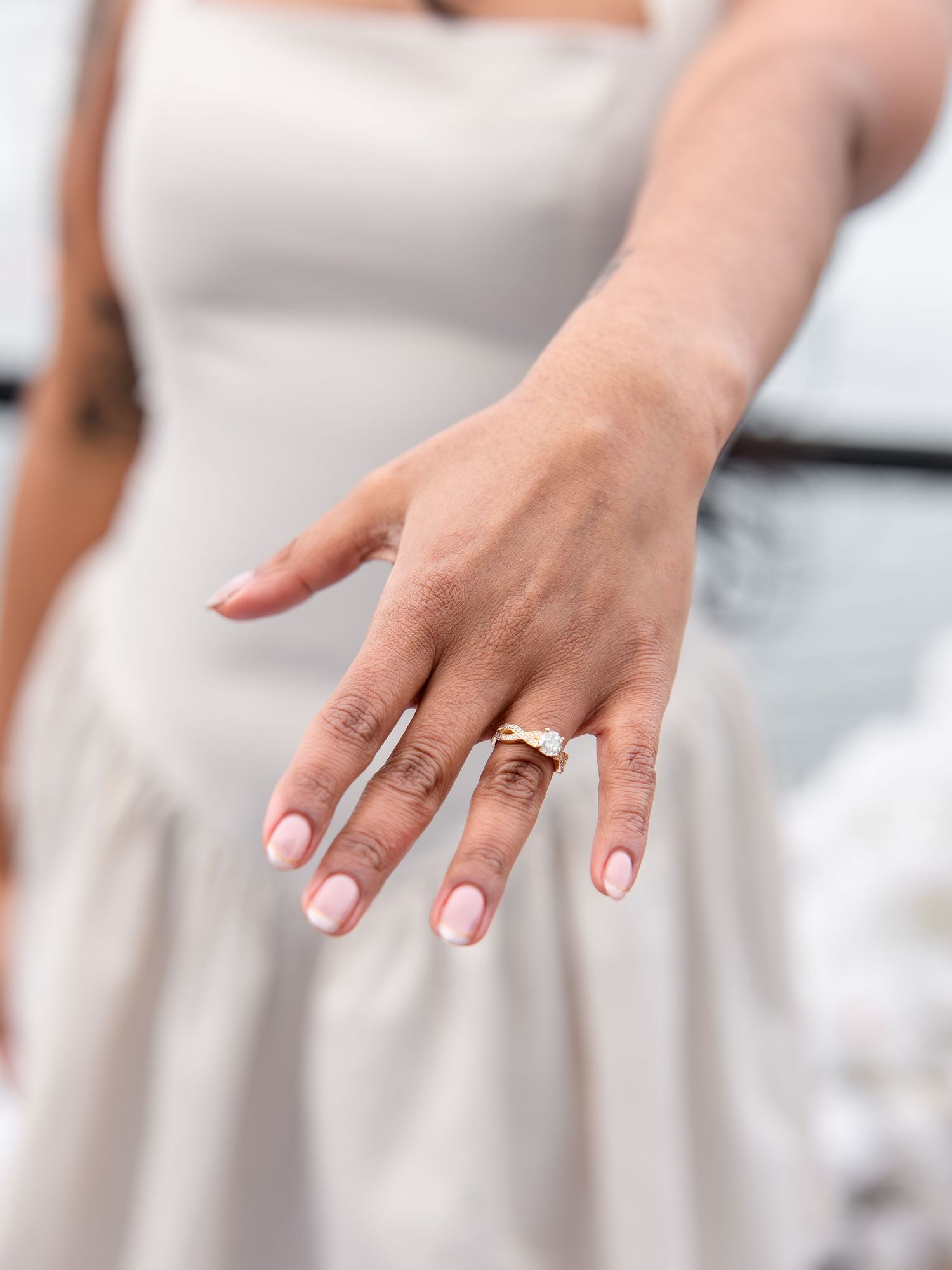 Woman showing off gold engagement ring wearing white dress outdoors.