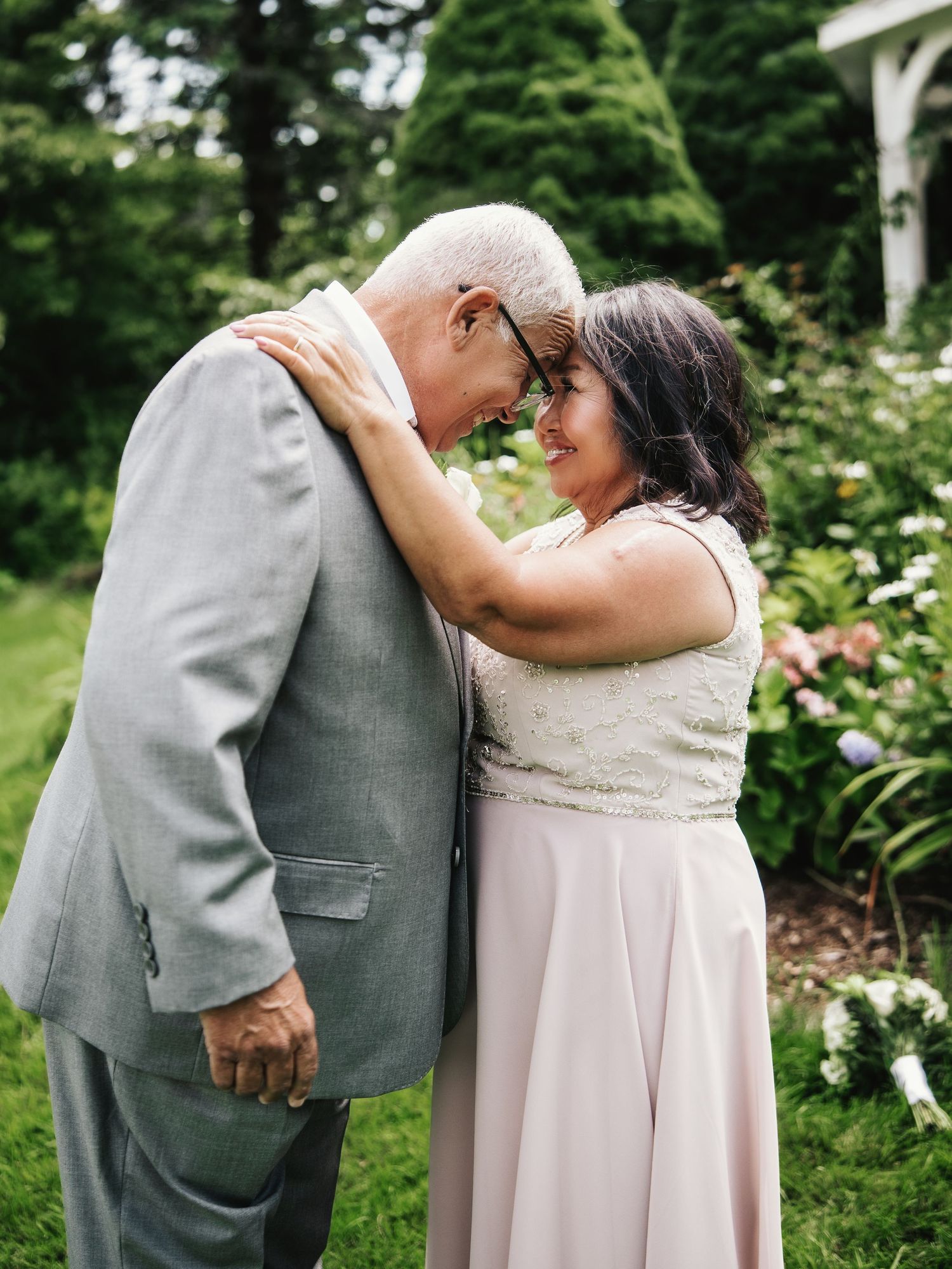 Elderly couple sharing a tender forehead-to-forehead moment in a garden, dressed in wedding attire.