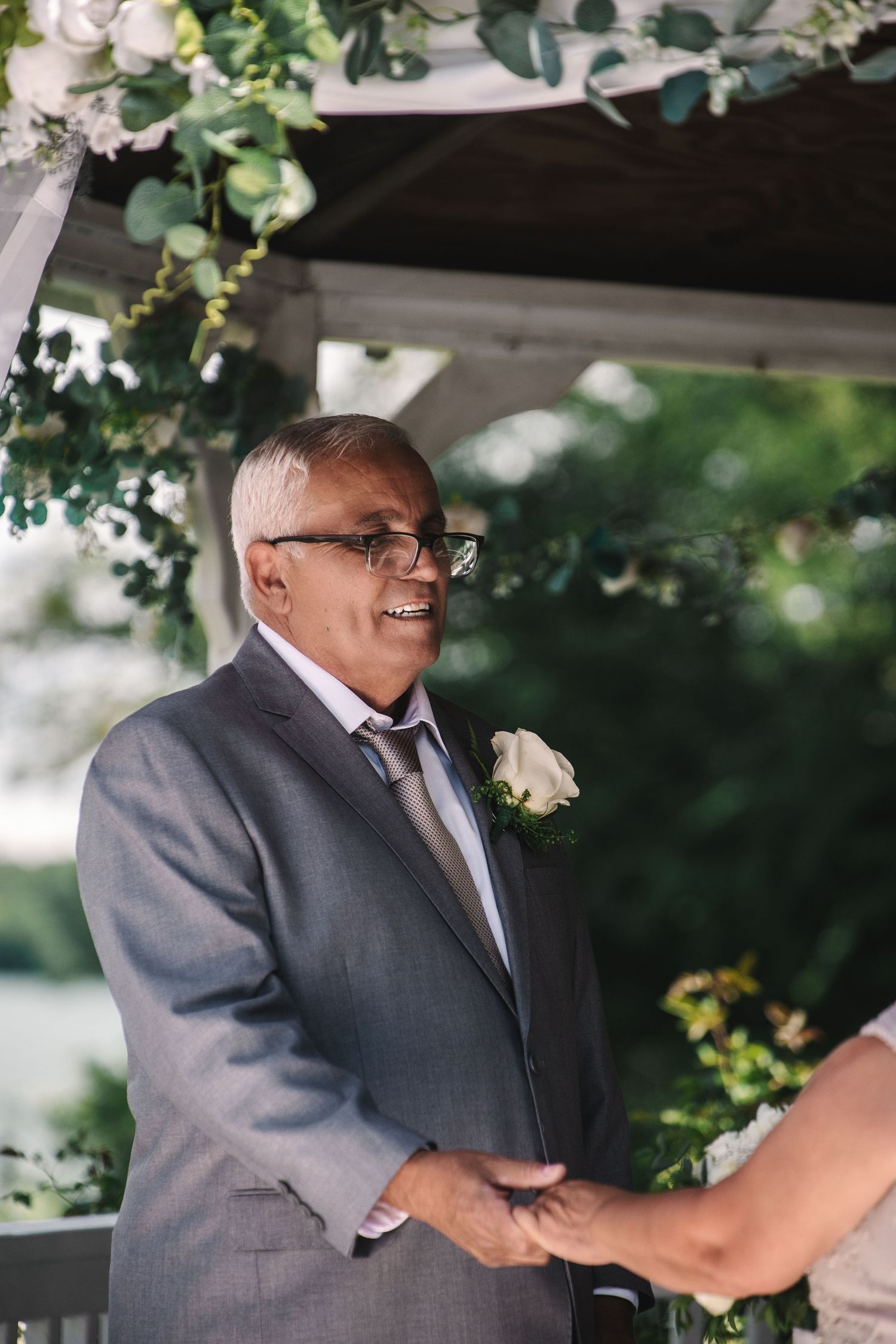 Smiling older man in gray suit with white rose boutonniere holding hands at outdoor wedding ceremony under floral arch.
