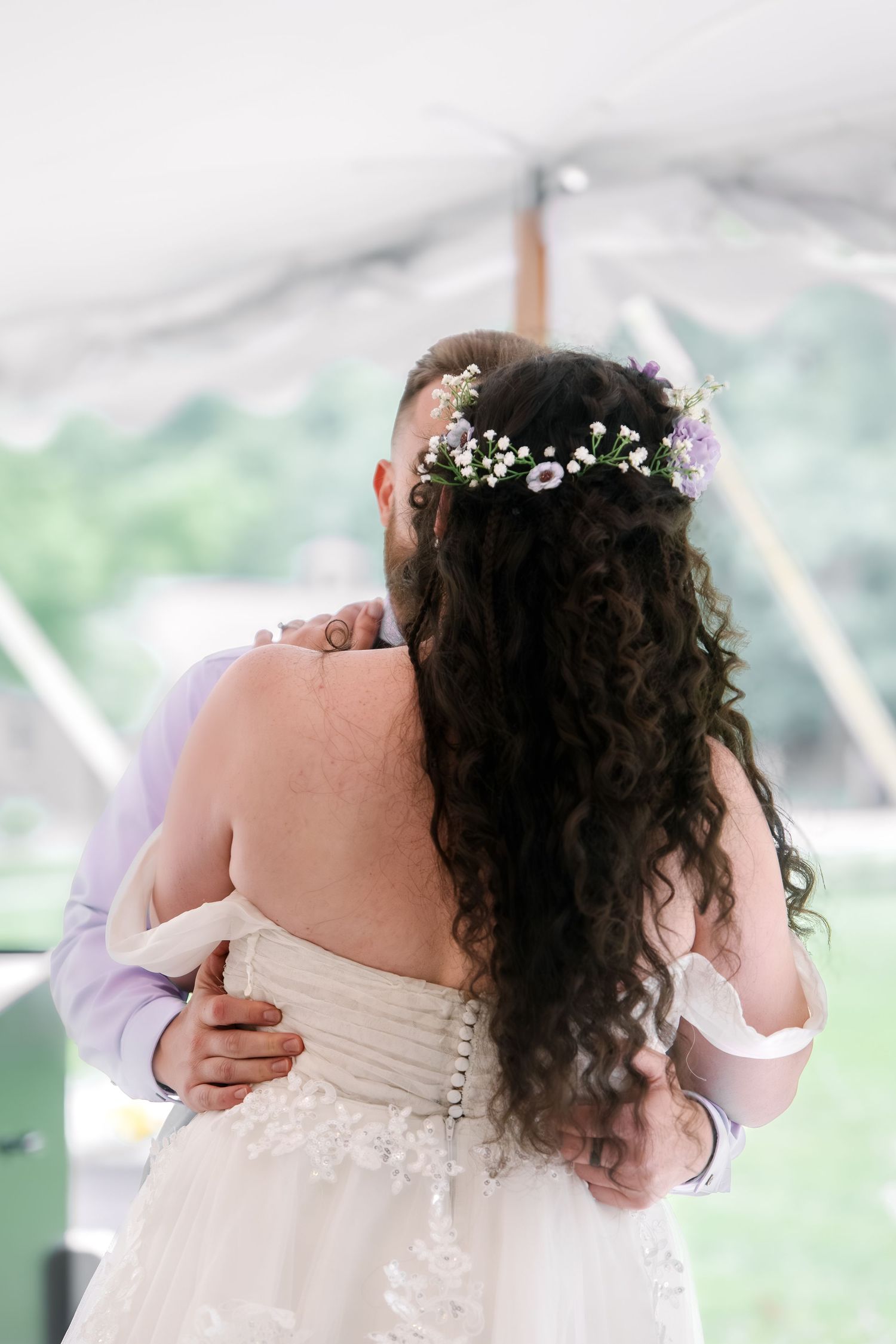 Bride and groom share first dance under tent, bride wearing floral crown and lace gown with curly hair.