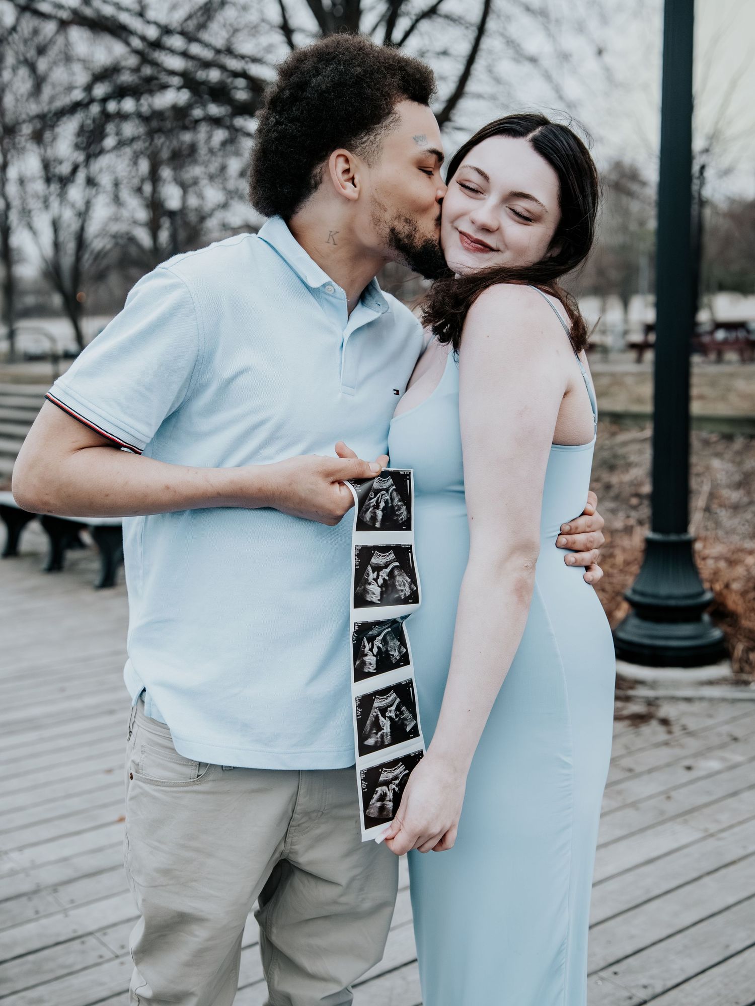 Happy couple holding ultrasound photos in a park, man kissing pregnant woman in matching light blue outfits.