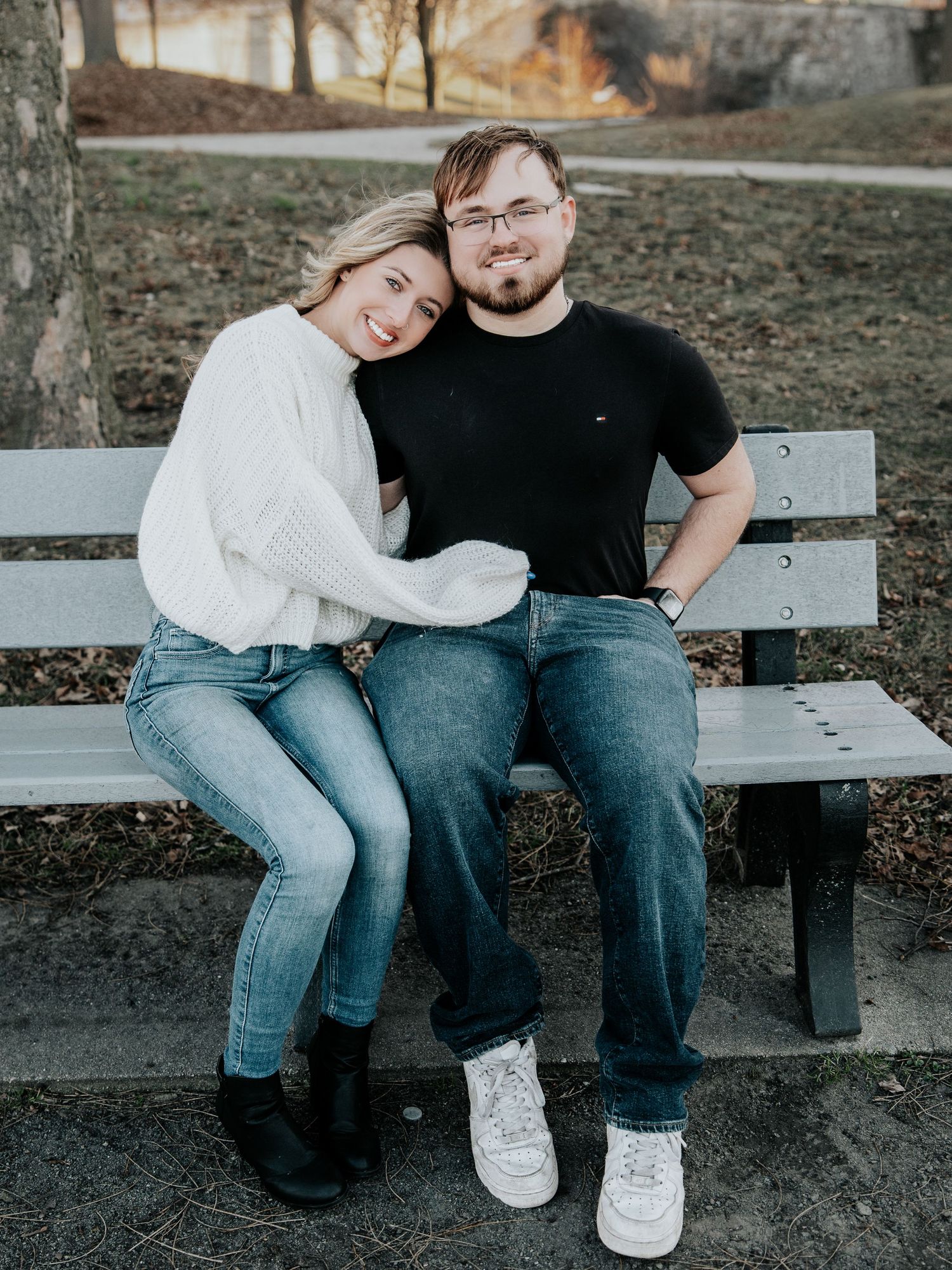 A smiling couple sits closely on a park bench, woman in white sweater and man in black shirt, both wearing jeans.