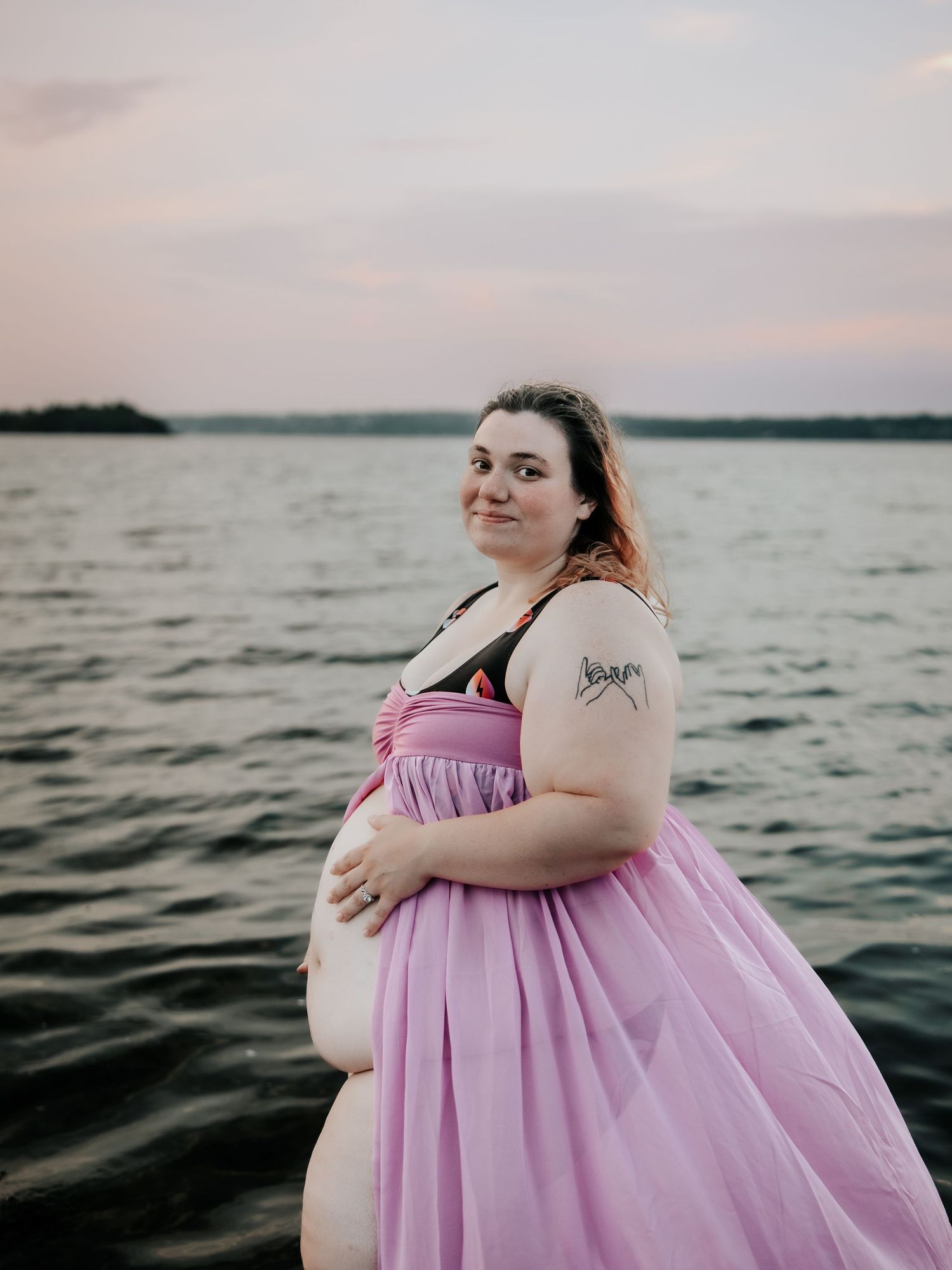 Pregnant woman in pink flowing skirt poses by a lake at sunset, showing baby bump with tattoo on arm.